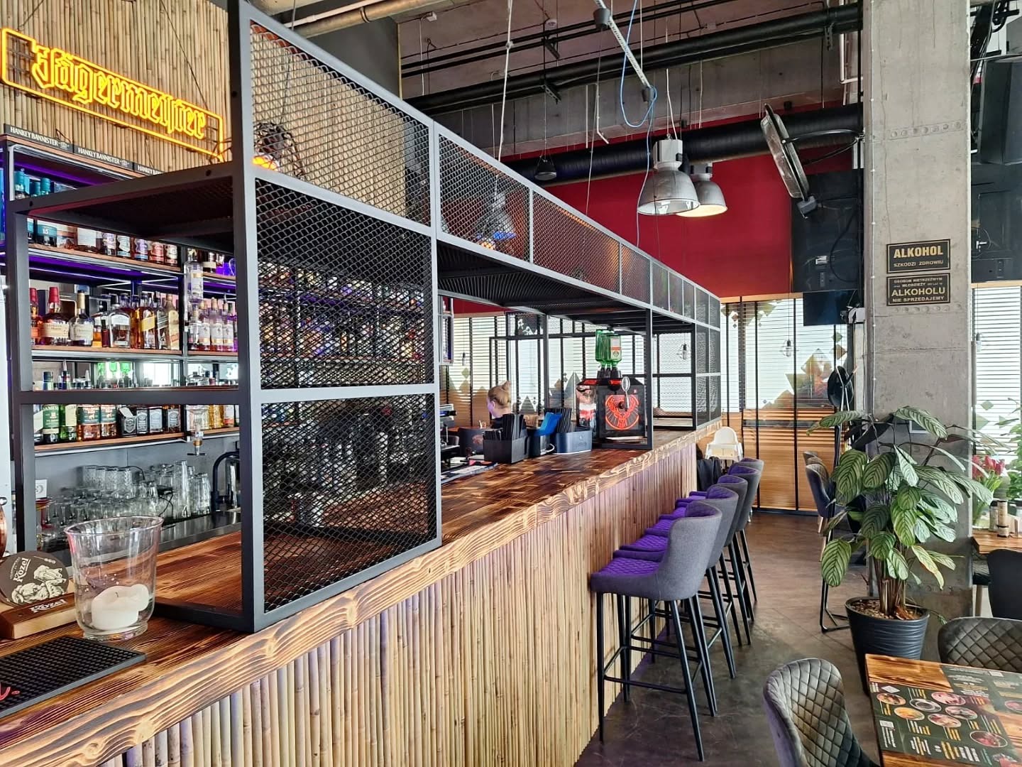 Bar w Rumi z drewnianym blatem i metalową konstrukcją nad barem. Widoczne butelki alkoholi i krzesła barowe z fioletowymi siedziskami, roślina doniczkowa.
