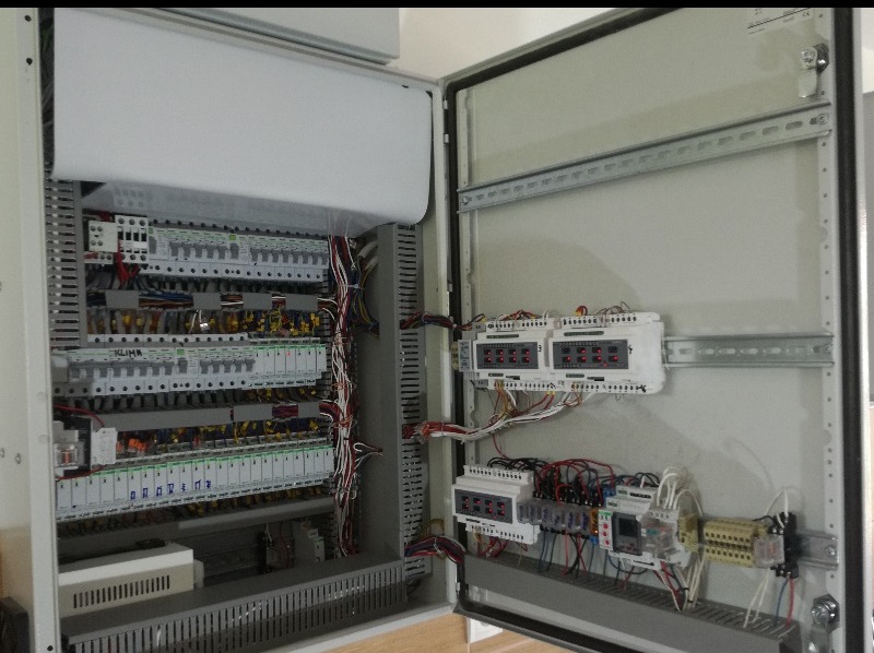 Otwarta szafa sterownicza z widocznymi elementami elektrycznymi, przewodami i modułami. Widoczne szyny montażowe i listwy zaciskowe.