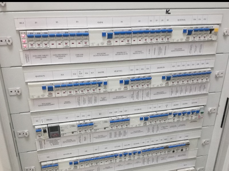 Rozdzielnia elektryczna z bezpiecznikami, opisana etykietami. Widoczne wyłączniki, zabezpieczenia i oznaczenia obwodów. Instalacja w kolorze białym.