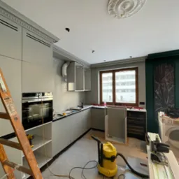 Montaż kuchni w trakcie realizacji w Warszawie. Widoczny odkurzacz, drabina i narzędzia. Elementy wykończeniowe sufitu dodają elegancji.