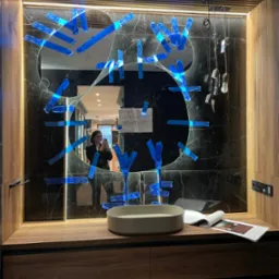 Nowoczesna łazienka z marmurową ścianą i okrągłym lustrem zabezpieczonym niebieską taśmą. Na blacie umywalka i otwarty magazyn wnętrzarski.