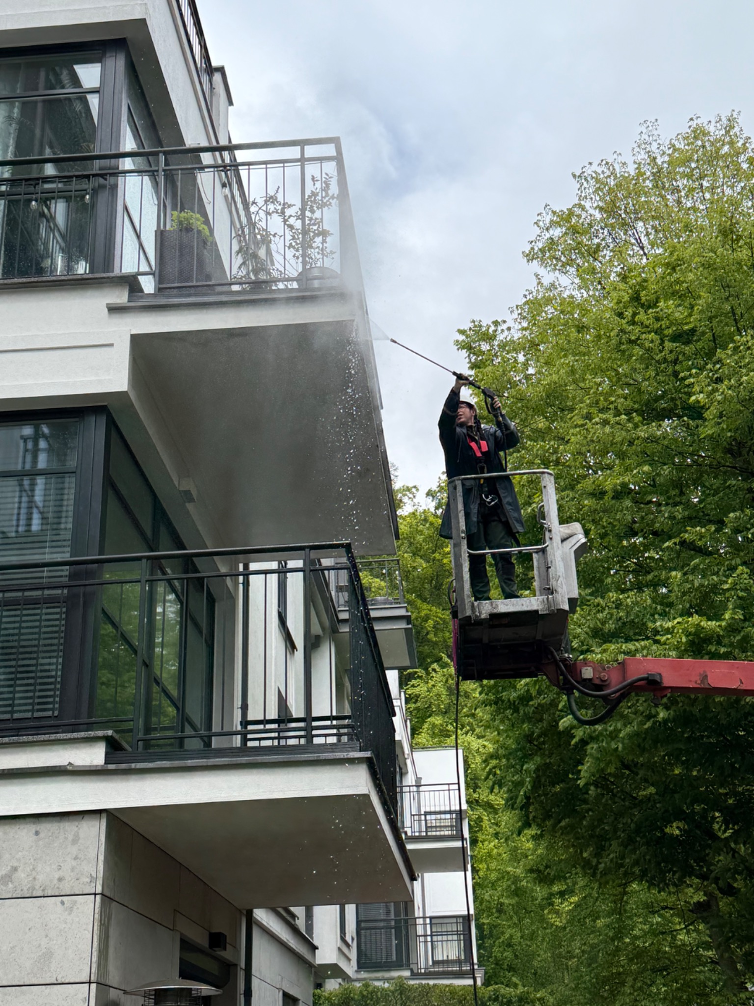 Pracownik na podnośniku czyści elewację balkonu myjką ciśnieniową. Widoczne ślady spływającej wody. W tle zieleń drzew.