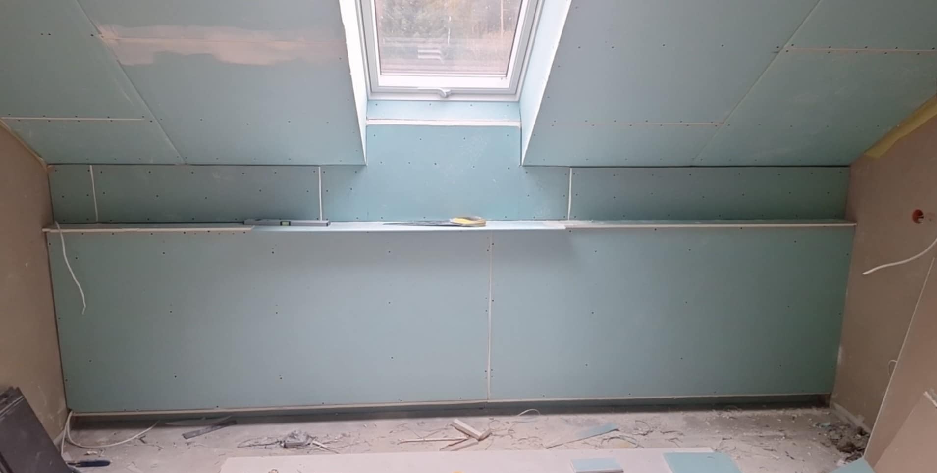 Poddasze z oknem dachowym w trakcie montażu płyt gipsowo-kartonowych. Widoczne narzędzia i poziomica. Ściany i sufit pokryte płytami, przygotowane do dalszych prac.