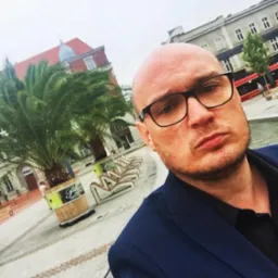 Mężczyzna w okularach i marynarce na tle placu miejskiego z palmami, budynkami i tramwajem w oddali, sfotografowany z bliska.