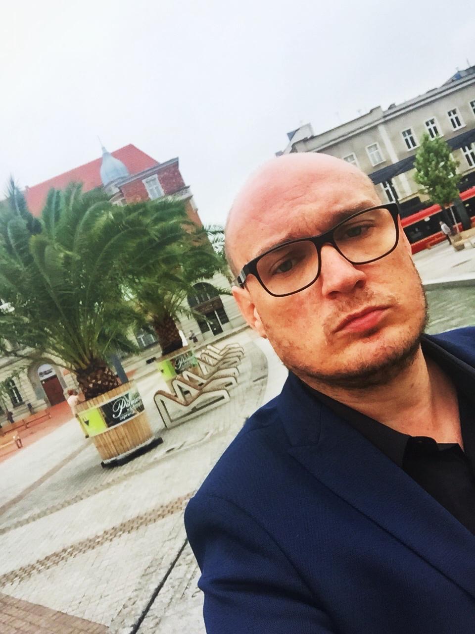 Mężczyzna w okularach i marynarce na tle placu miejskiego z palmami, budynkami i tramwajem w oddali, sfotografowany z bliska.