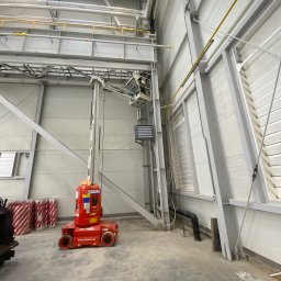 NEON - Prace na wysokości w hali przemysłowej: pracownik na podnośniku montuje instalacje pod sufitem. Widoczne rury, kable i konstrukcja hali.