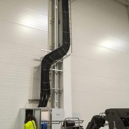 NEON - Montaż instalacji elektrycznej w hali: pracownik w żółtej kamizelce łączy kable w szafie, widoczny podnośnik i koryto kablowe prowadzące do góry.