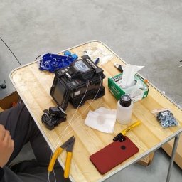 NEON - Fuzja światłowodów na stole: spawarka światłowodowa, obcinak, stripper, telefon. Widoczny etap pracy, elementy instalacji i narzędzia.