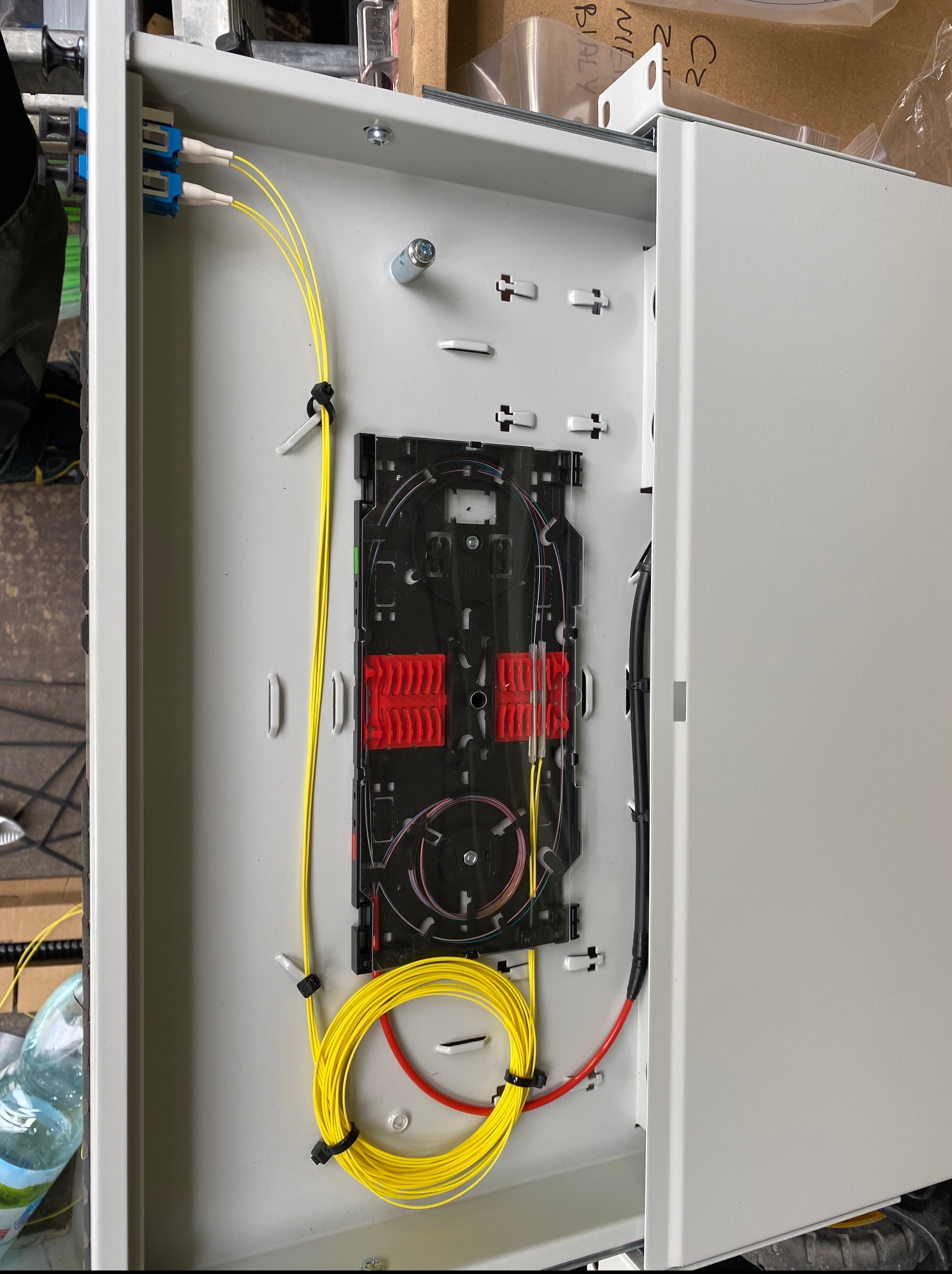 Otwarta, biała skrzynka z zamontowanym panelem światłowodowym, widoczne żółte kable światłowodowe i czarne kable w tle. Precyzyjne ułożenie kabli.