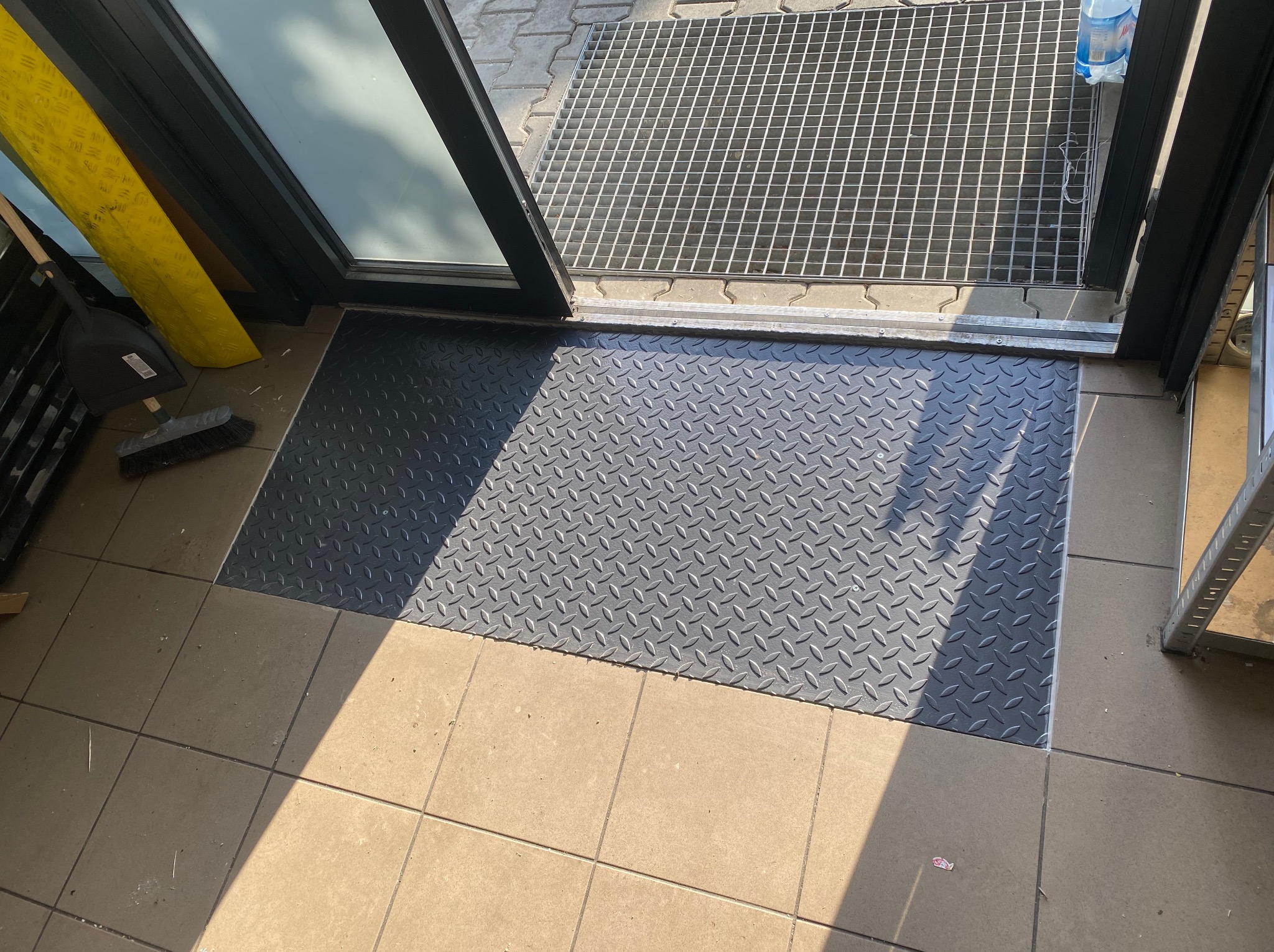 Czarna, ryflowana mata wejściowa wkomponowana w płytki podłogowe przy wejściu z metalową kratą, częściowo widoczna żółta osłona kabli i szczotka.