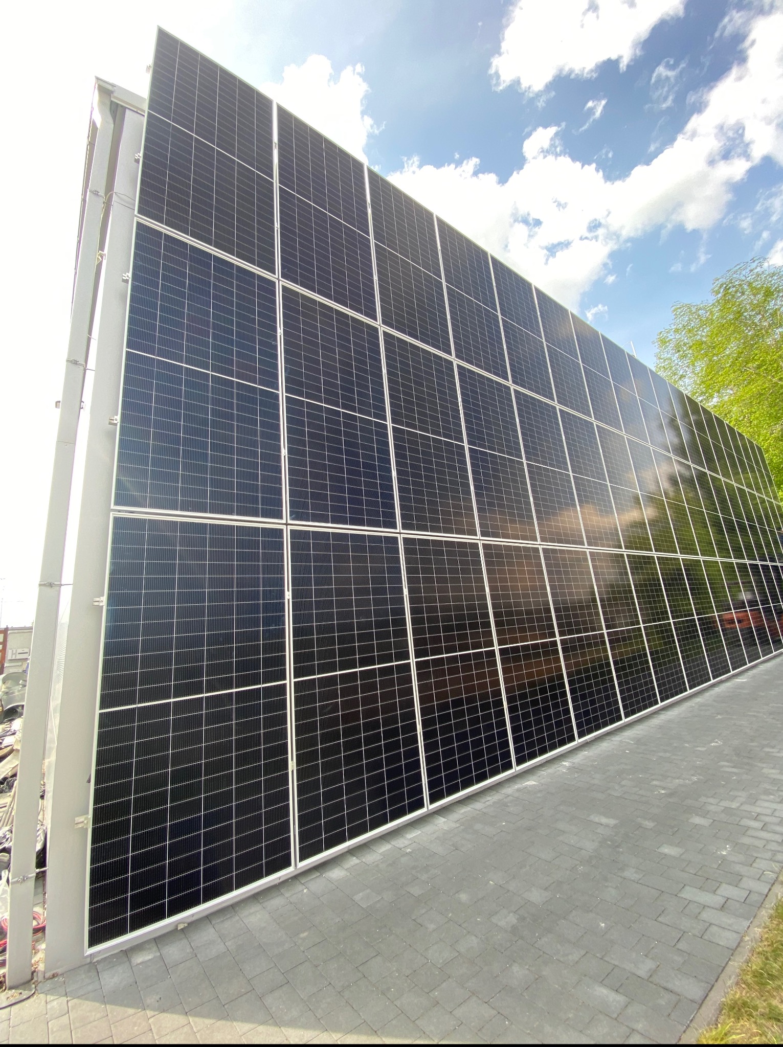 Duża instalacja fotowoltaiczna na szarej konstrukcji, widok z dołu, błękitne niebo z chmurami w tle. Panele odbijają światło, leżąc na szarym chodniku.