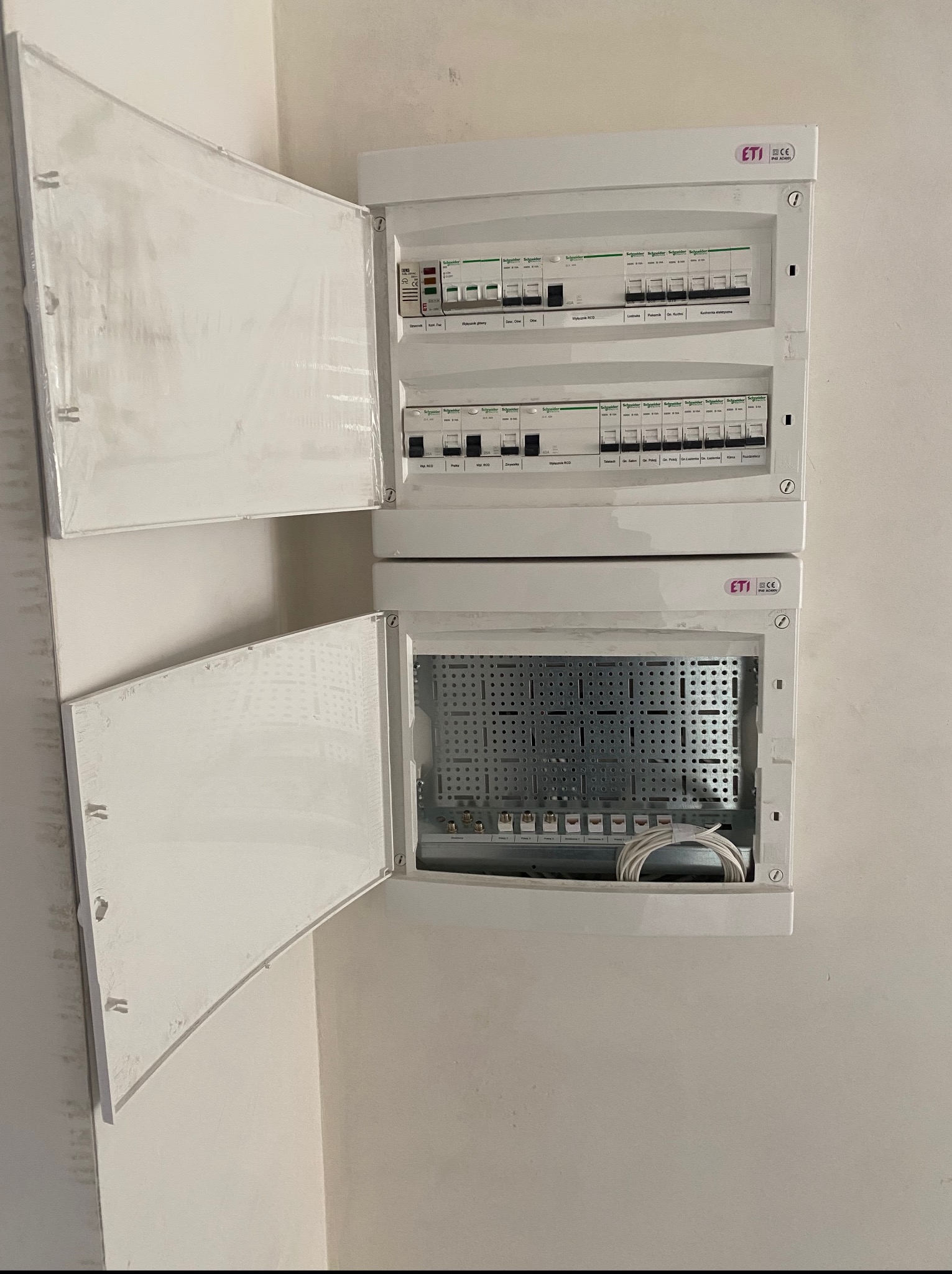 Dwa białe panele elektryczne ETI zamontowane na ścianie, jeden z otwartymi drzwiczkami, drugi z widocznymi bezpiecznikami Schneider. Widoczne przewody.