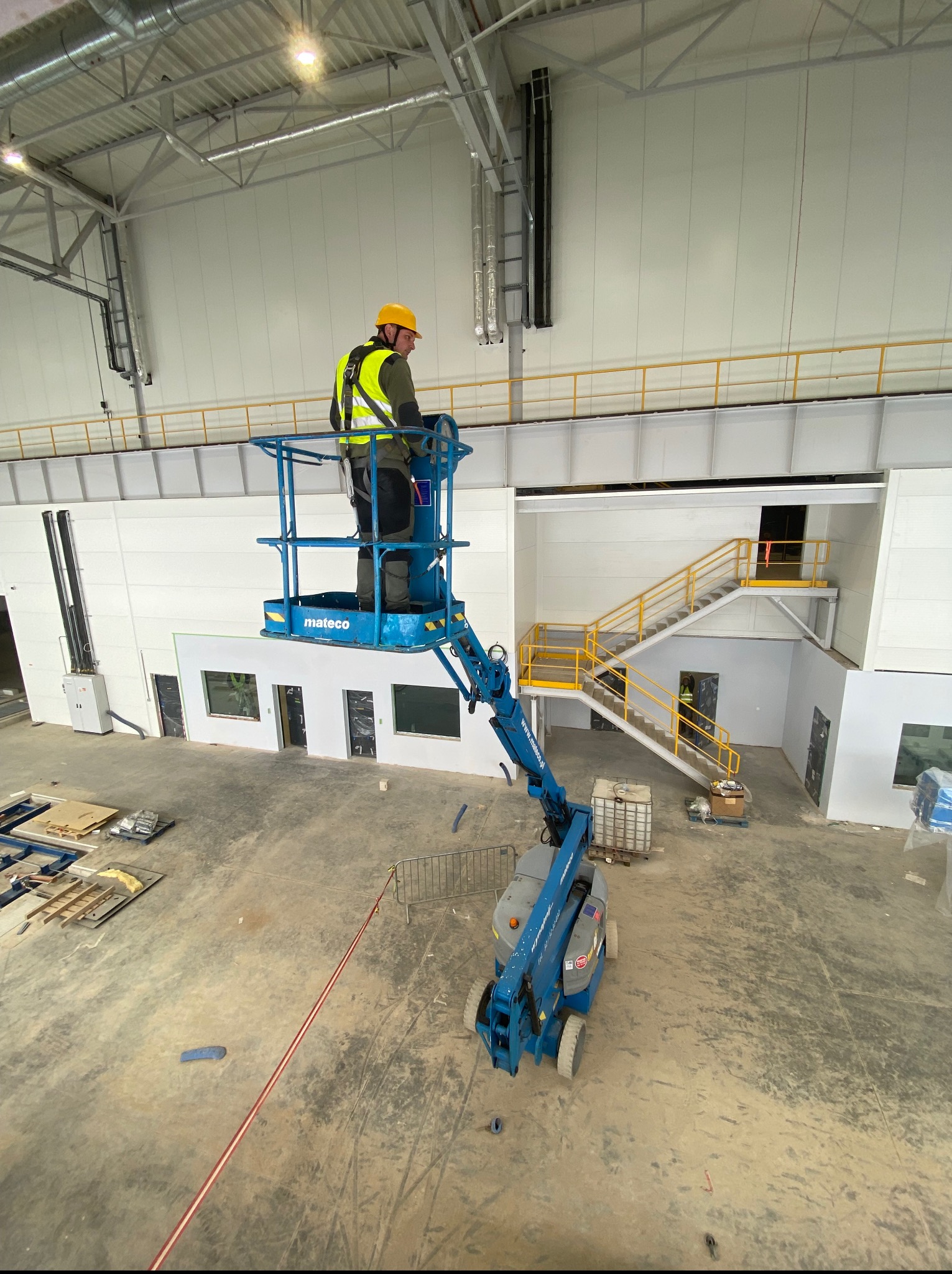 Pracownik w żółtym kasku na podnośniku koszowym w hali przemysłowej. Widoczne instalacje pod sufitem i częściowo wykończone ściany.