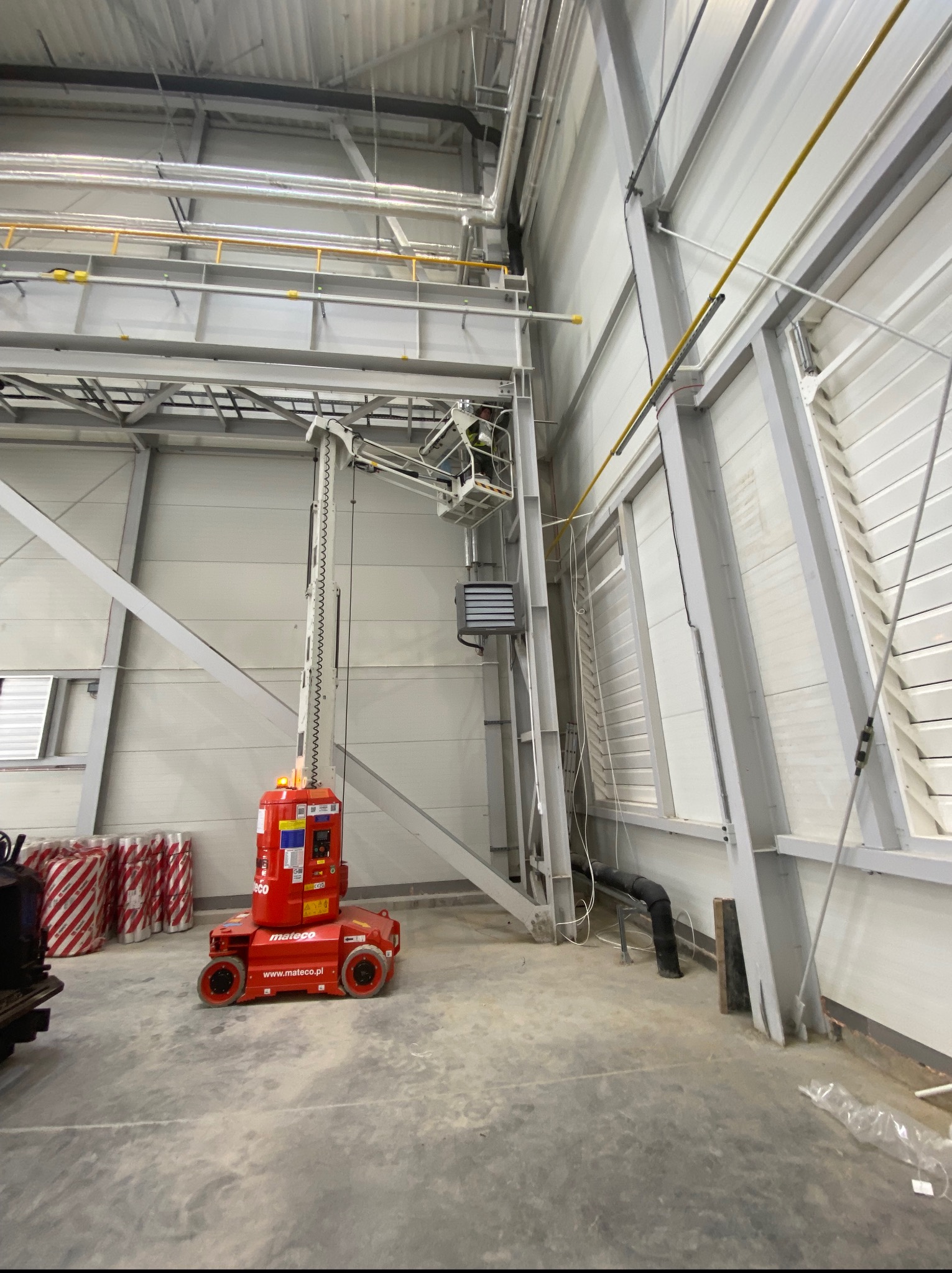 Prace na wysokości w hali przemysłowej: pracownik na podnośniku montuje instalacje pod sufitem. Widoczne rury, kable i konstrukcja hali.