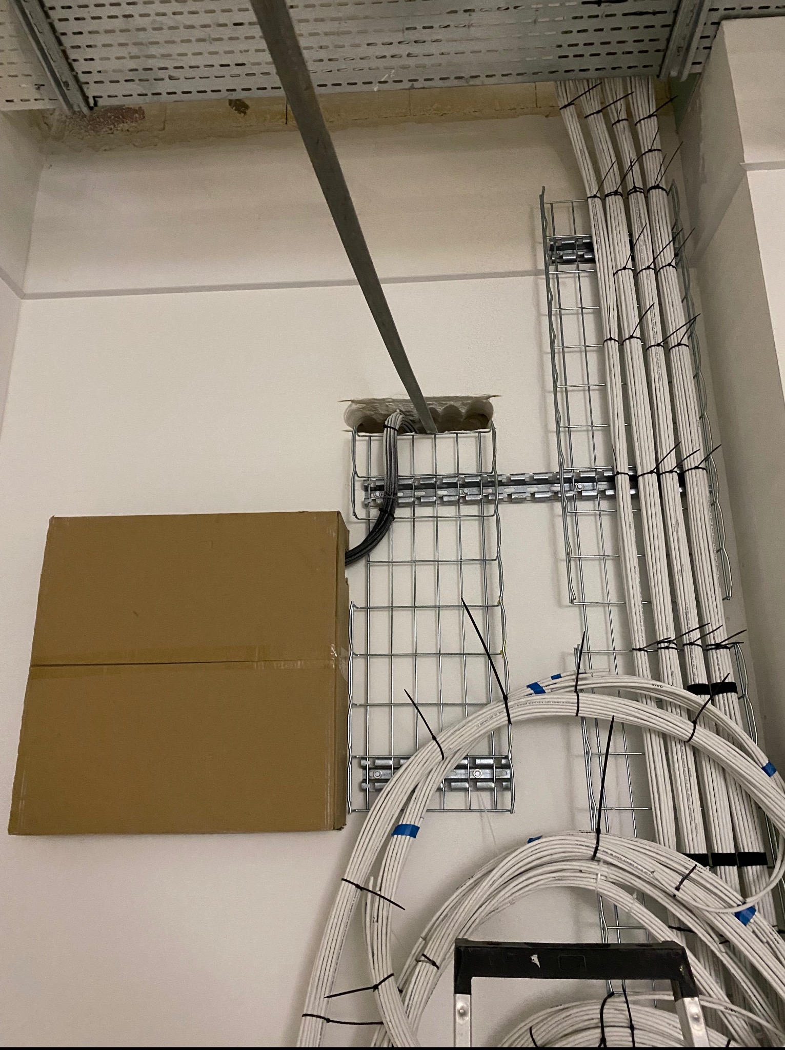 Białe kable ułożone w metalowych korytach na ścianie, zabezpieczone opaskami zaciskowymi. Obok kartonowe pudło i drabina. Widoczne elementy instalacji pod sufitem.
