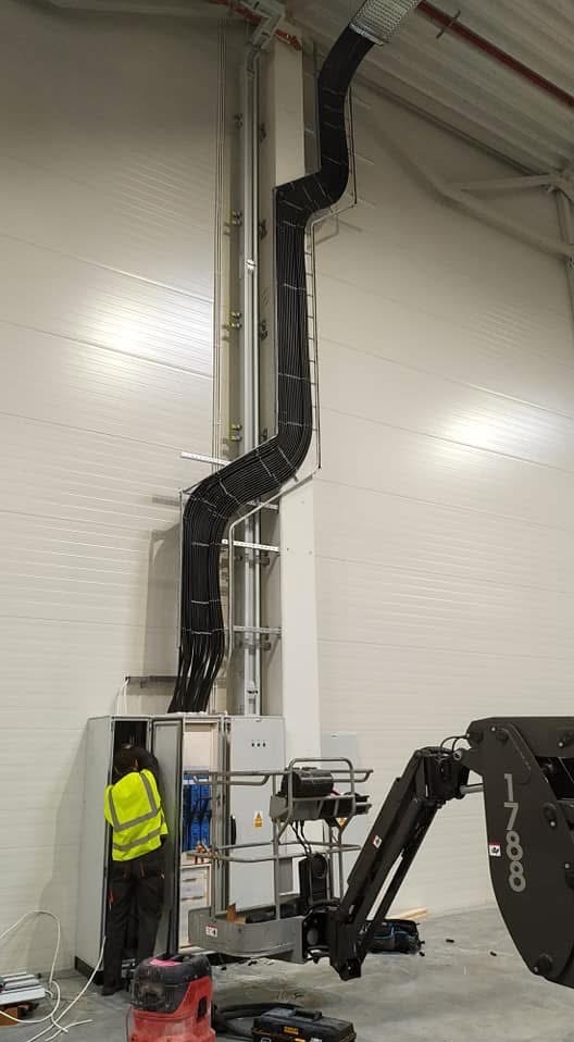 Montaż instalacji elektrycznej w hali: pracownik w żółtej kamizelce łączy kable w szafie, widoczny podnośnik i koryto kablowe prowadzące do góry.