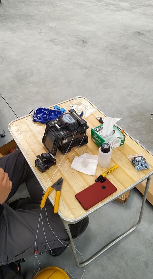 Fuzja światłowodów na stole: spawarka światłowodowa, obcinak, stripper, telefon. Widoczny etap pracy, elementy instalacji i narzędzia.