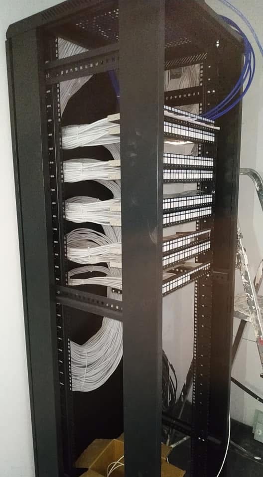 Czarna szafa rack z panelami krosowniczymi i uporządkowanymi kablami w kolorze białym i niebieskim, widoczne drabiny kablowe i fragment drabiny malarskiej.