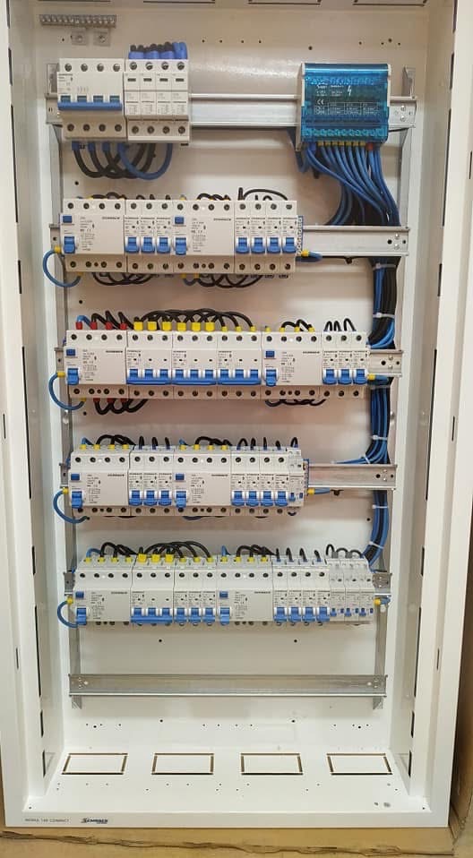 Rozdzielnia elektryczna z licznymi zabezpieczeniami, starannie ułożone przewody w kolorze niebieskim i czarnym, widok z przodu na białą obudowę.