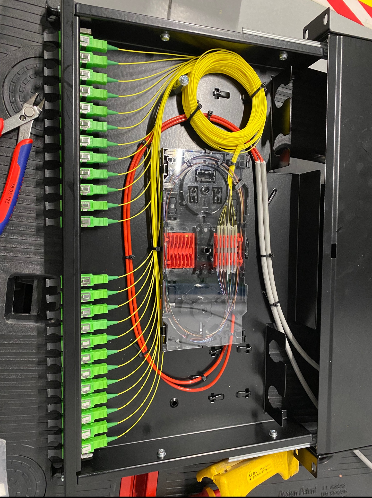 Panel światłowodowy z precyzyjnie ułożonymi kablami w kolorach żółtym, czerwonym i szarym. Widoczne złącza światłowodowe z zielonymi końcówkami. Użyte narzędzia: obcinaczki.