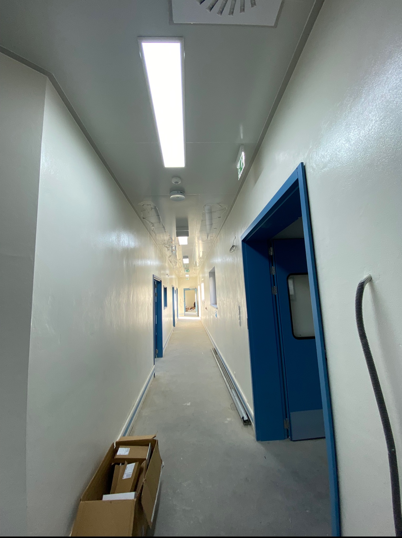 Długi, jasny korytarz z białymi ścianami i niebieskimi drzwiami. Widoczne lampy sufitowe i kartonowe pudła na podłodze. Perspektywa zbieżna w głąb.