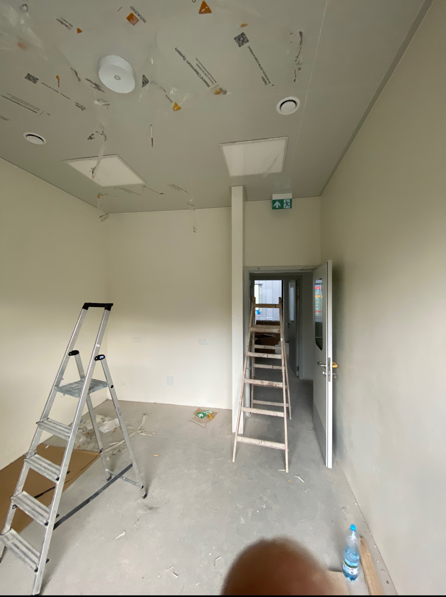 Wnętrze w trakcie wykończenia: dwie drabiny, sufit z zamontowanym oświetleniem, ślady po folii ochronnej, butelka wody. Widoczny korytarz z otwartymi drzwiami.