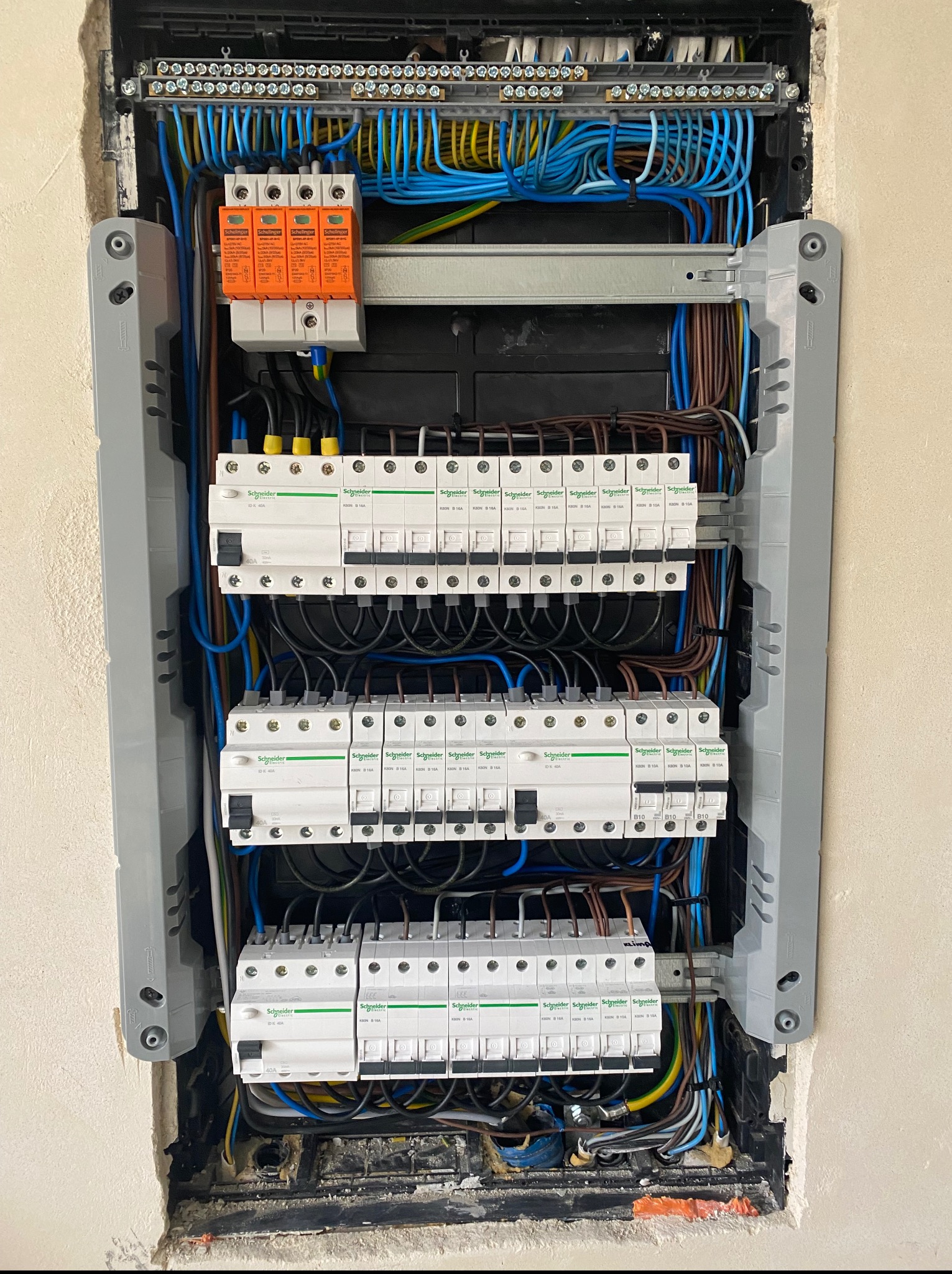 Rozdzielnia elektryczna Schneider z zabezpieczeniami przepięciowymi i wyłącznikami, widoczna wnęka w ścianie, instalacja w trakcie montażu.