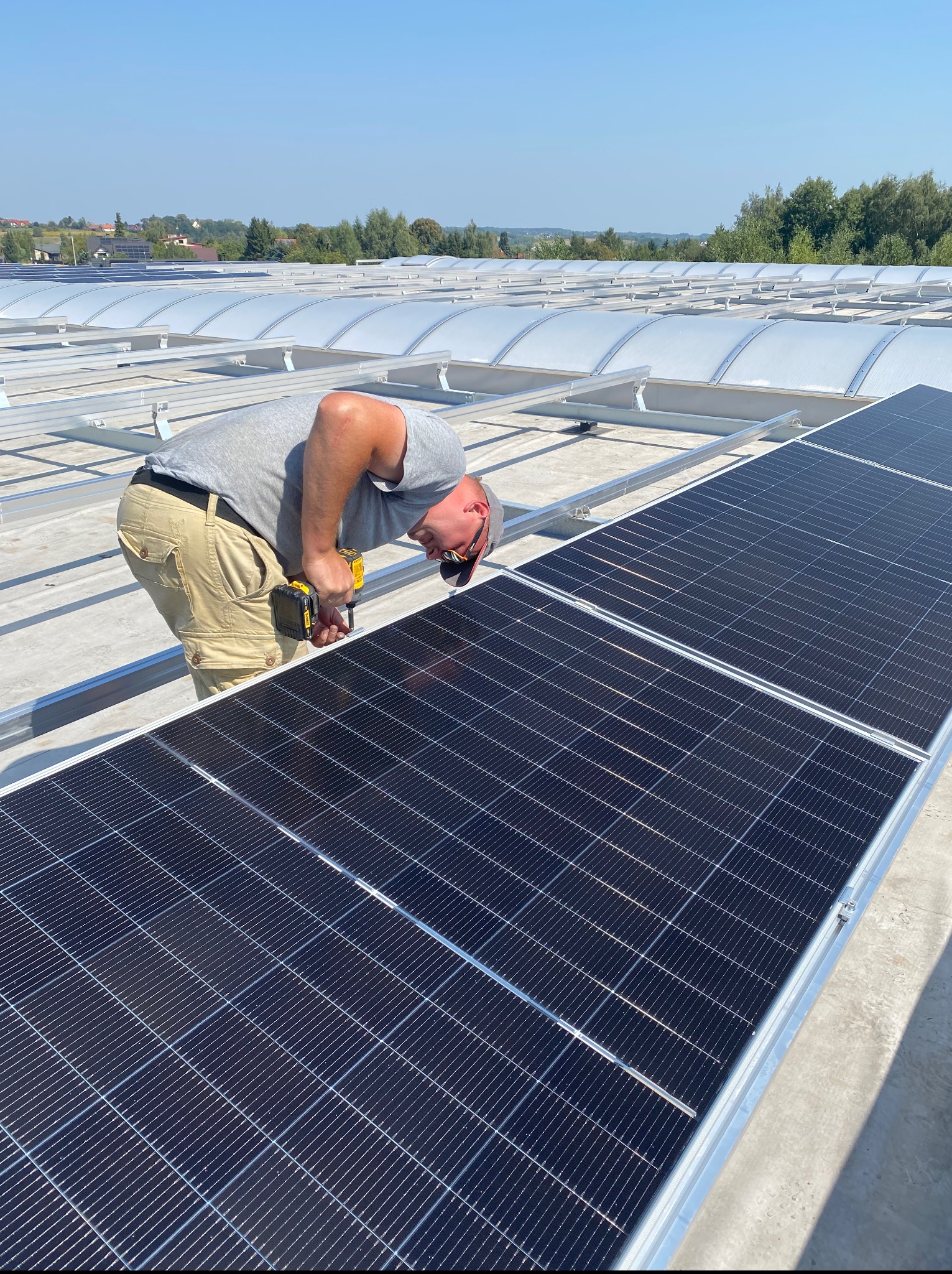 Mężczyzna montuje panele słoneczne na dachu budynku przemysłowego w słoneczny dzień. Widoczne narzędzia i elementy konstrukcji.