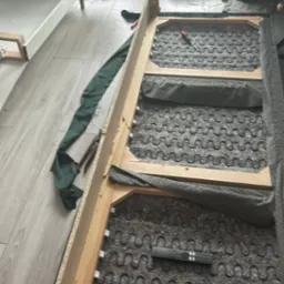 Demontaż sofy z zieloną tapicerką w jasnym wnętrzu. Widoczne sprężyny, drewniana rama i narzędzia. W tle okno balkonowe i choinka.