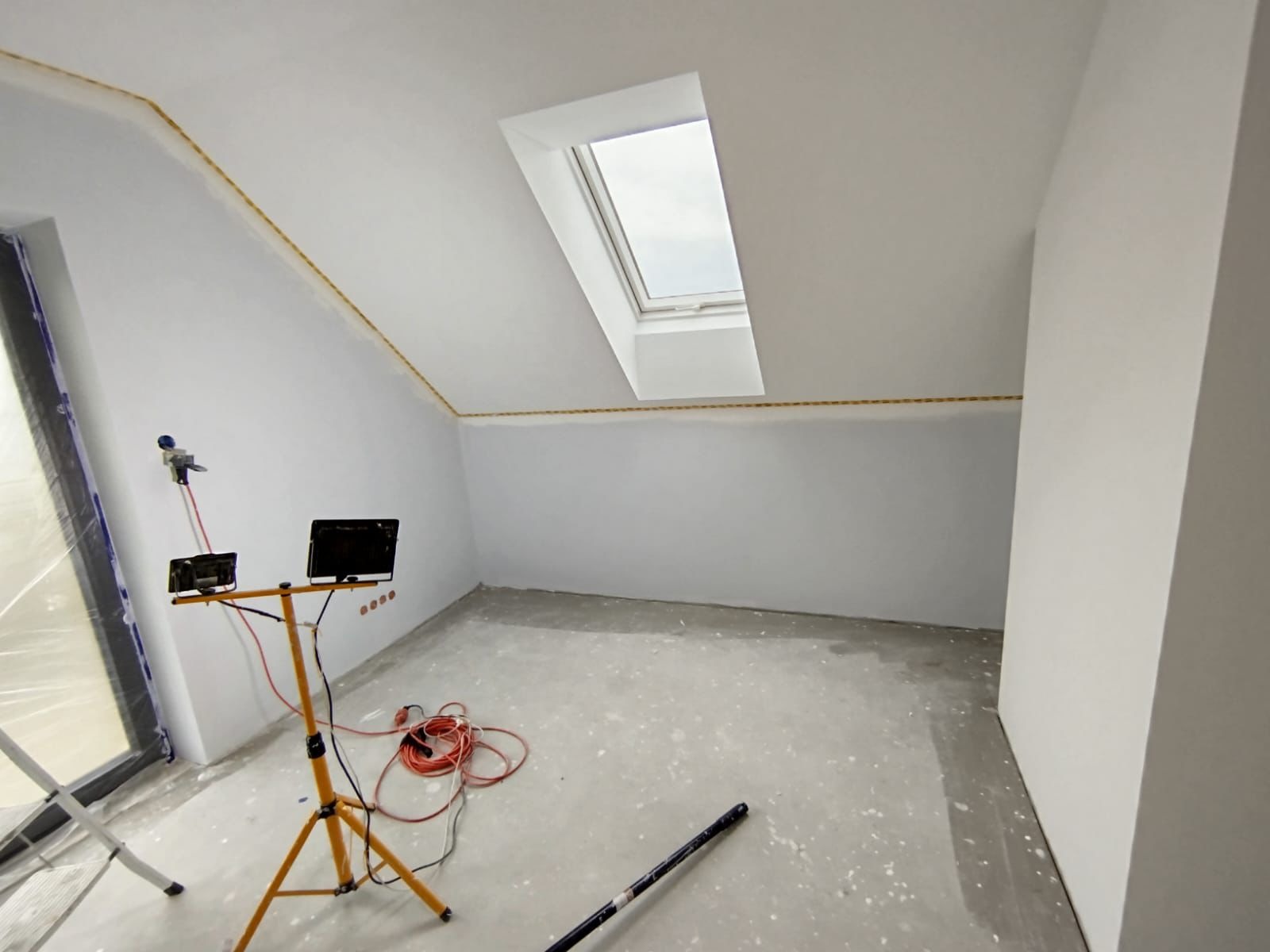 Pokój na poddaszu w trakcie malowania. Widoczne narzędzia malarskie, okno dachowe i zabezpieczone folią okno. Ściany przygotowane do prac wykończeniowych.