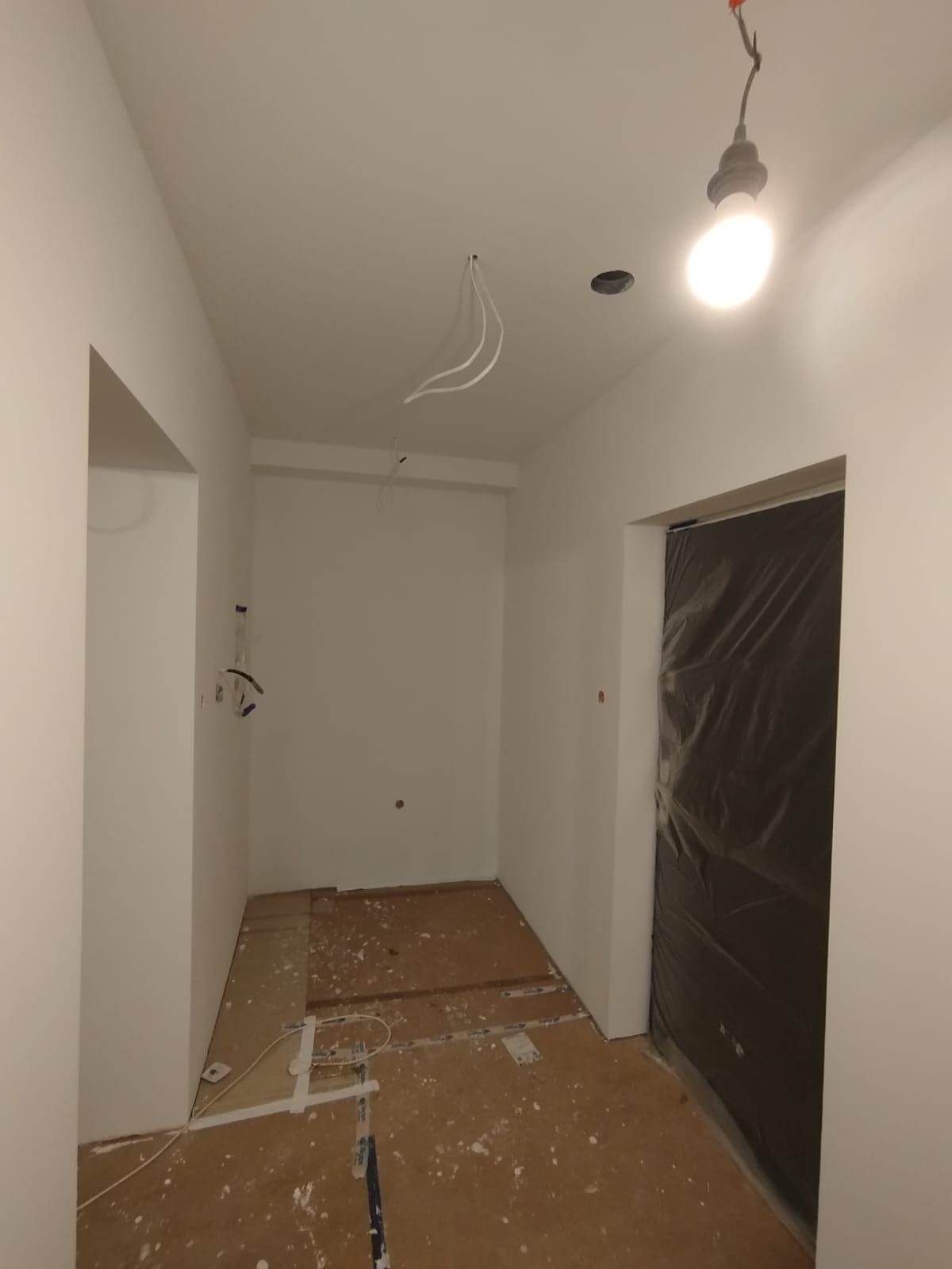 Wnętrze w trakcie remontu: białe ściany, zabezpieczone folią drzwi, widoczne przewody elektryczne i przygotowane podłoże pod panele. Robocze oświetlenie.