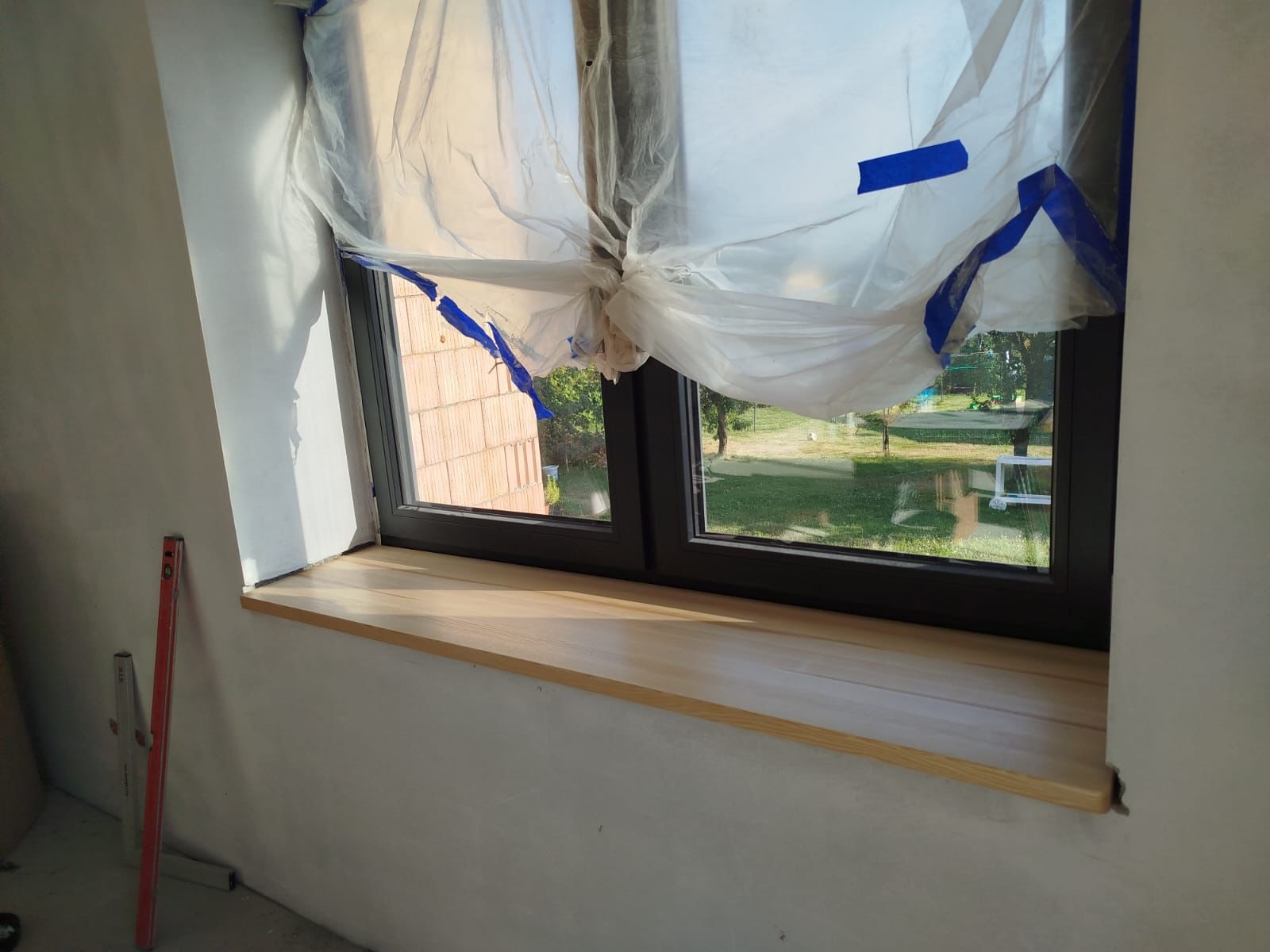 Nowy, drewniany parapet osadzony w oknie. Okno zabezpieczone folią malarską. Poziomica oparta o ścianę. Widok z okna na zieleń.