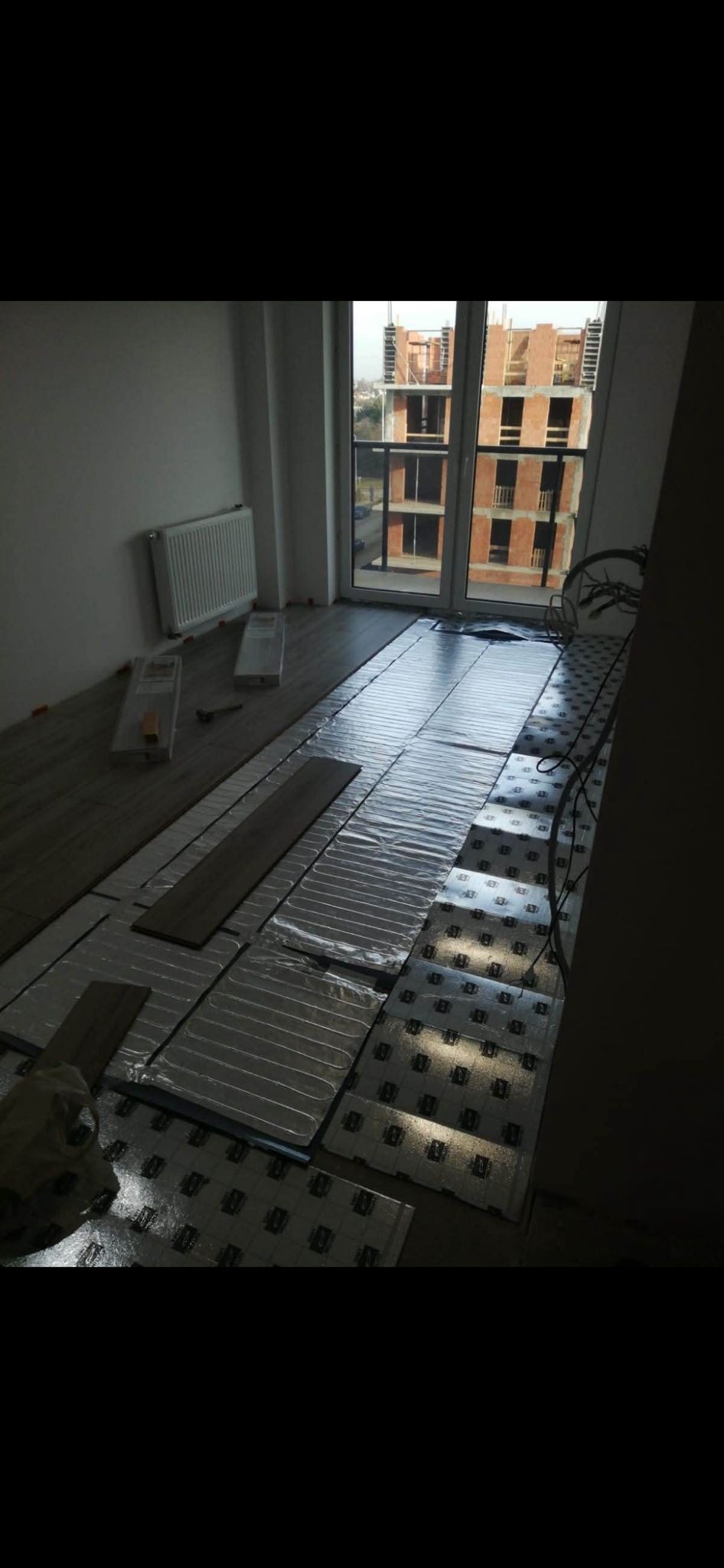 Wnętrze z częściowo ułożoną folią grzewczą pod panele, widoczne panele, grzejnik i budynek w budowie za oknem. Etap prac wykończeniowych w nowym mieszkaniu.