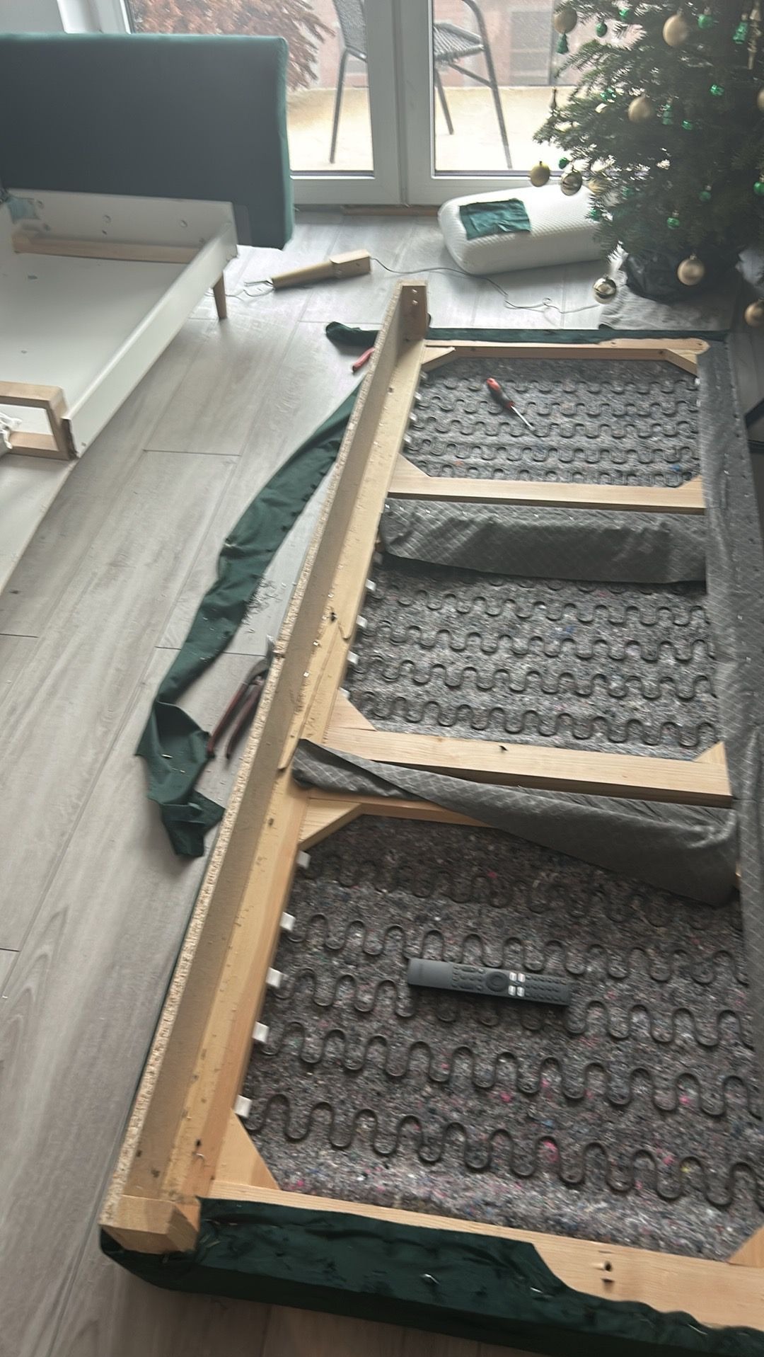 Demontaż sofy z zieloną tapicerką w jasnym wnętrzu. Widoczne sprężyny, drewniana rama i narzędzia. W tle okno balkonowe i choinka.