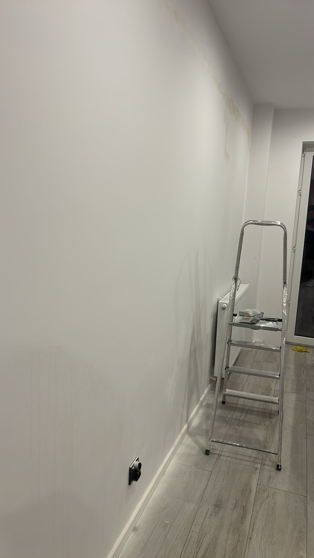 Biała ściana z zaciekami, drabina malarska, gniazdko elektryczne. Widoczne przygotowania do malowania ścian wewnątrz pomieszczenia.