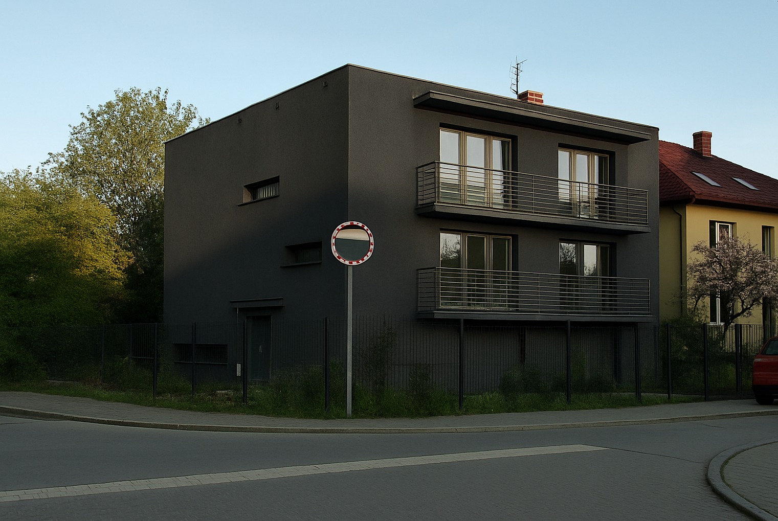 Dwukondygnacyjny, ciemnoszary dom z balkonami i płaskim dachem, widziany z rogu ulicy, obok znak drogowy. Nowoczesny design, minimalistyczna bryła, kontrast z zielenią.