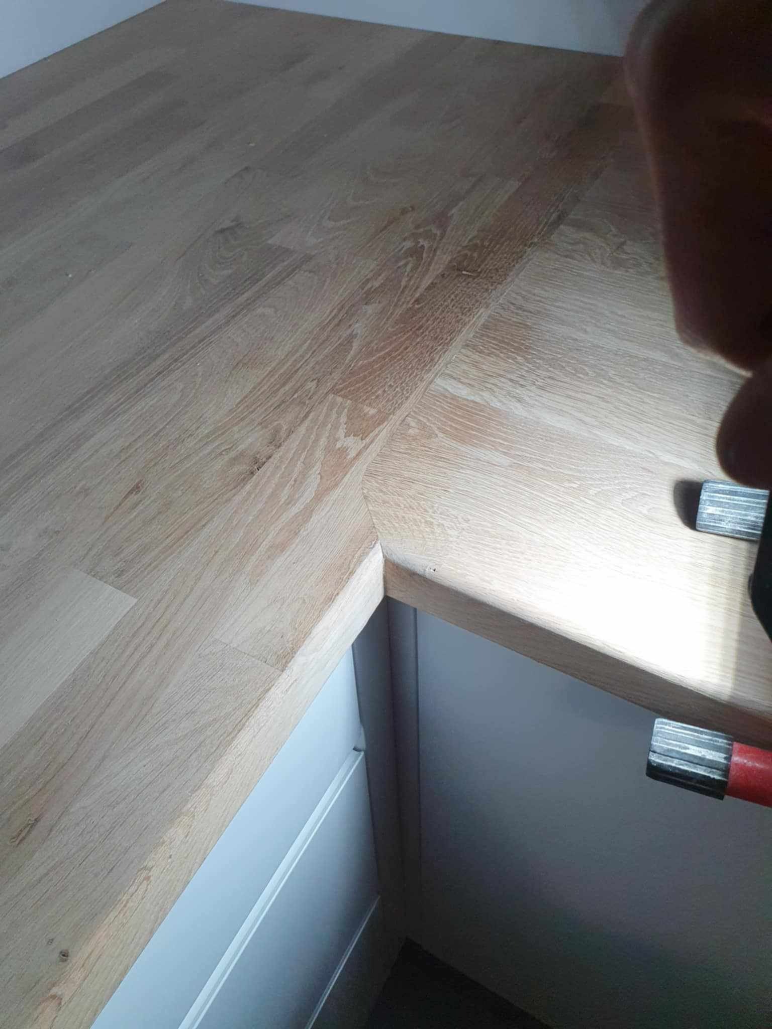 Blat drewniany, łączony pod kątem prostym, zamocowany do szafki. Widoczne ściski stolarskie. Jasne drewno, minimalistyczny design.