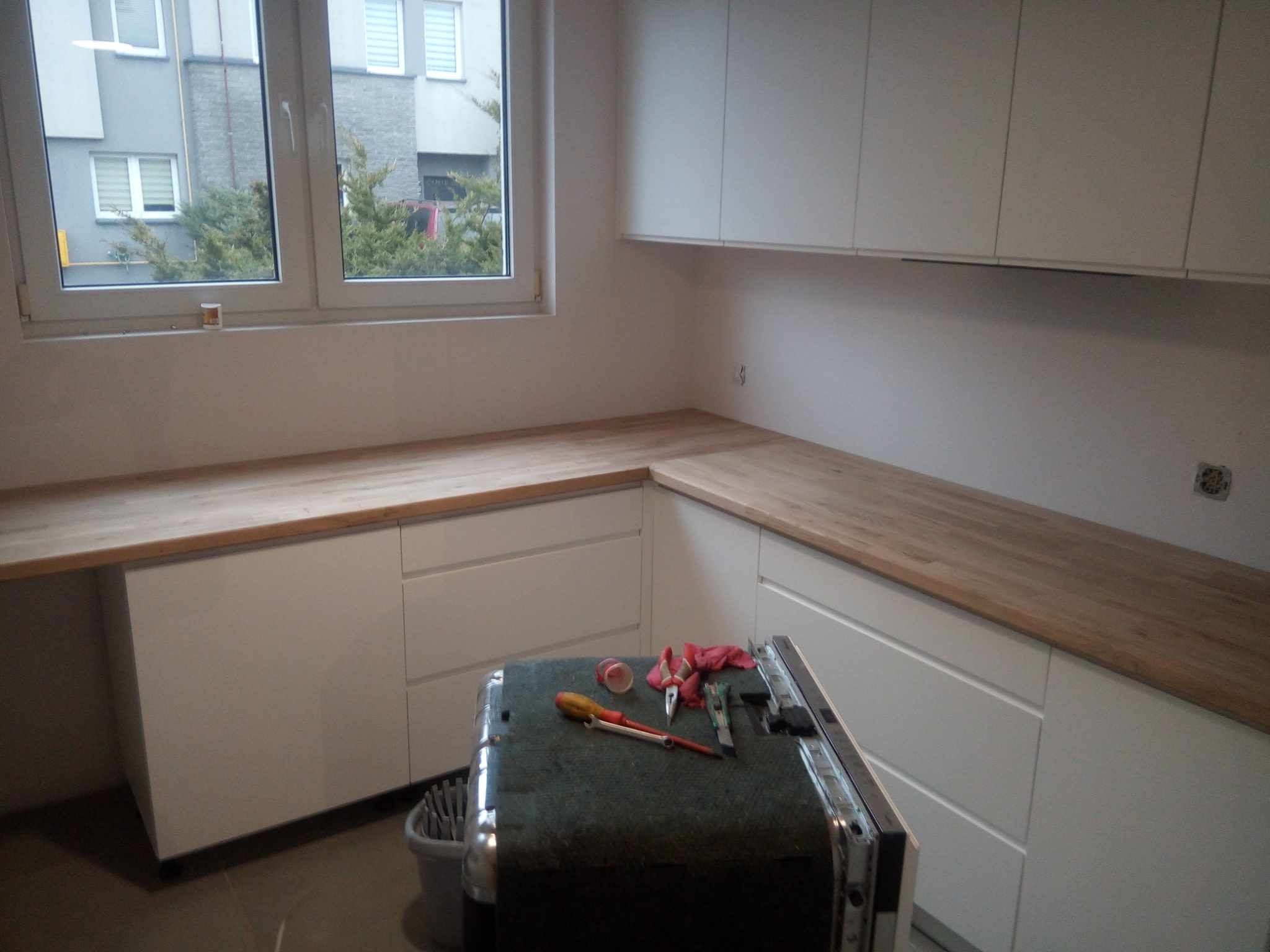 Montaż białej zabudowy kuchennej z drewnianym blatem. Widoczne narzędzia i niedokończona instalacja sprzętu AGD. Okno z widokiem na blok.
