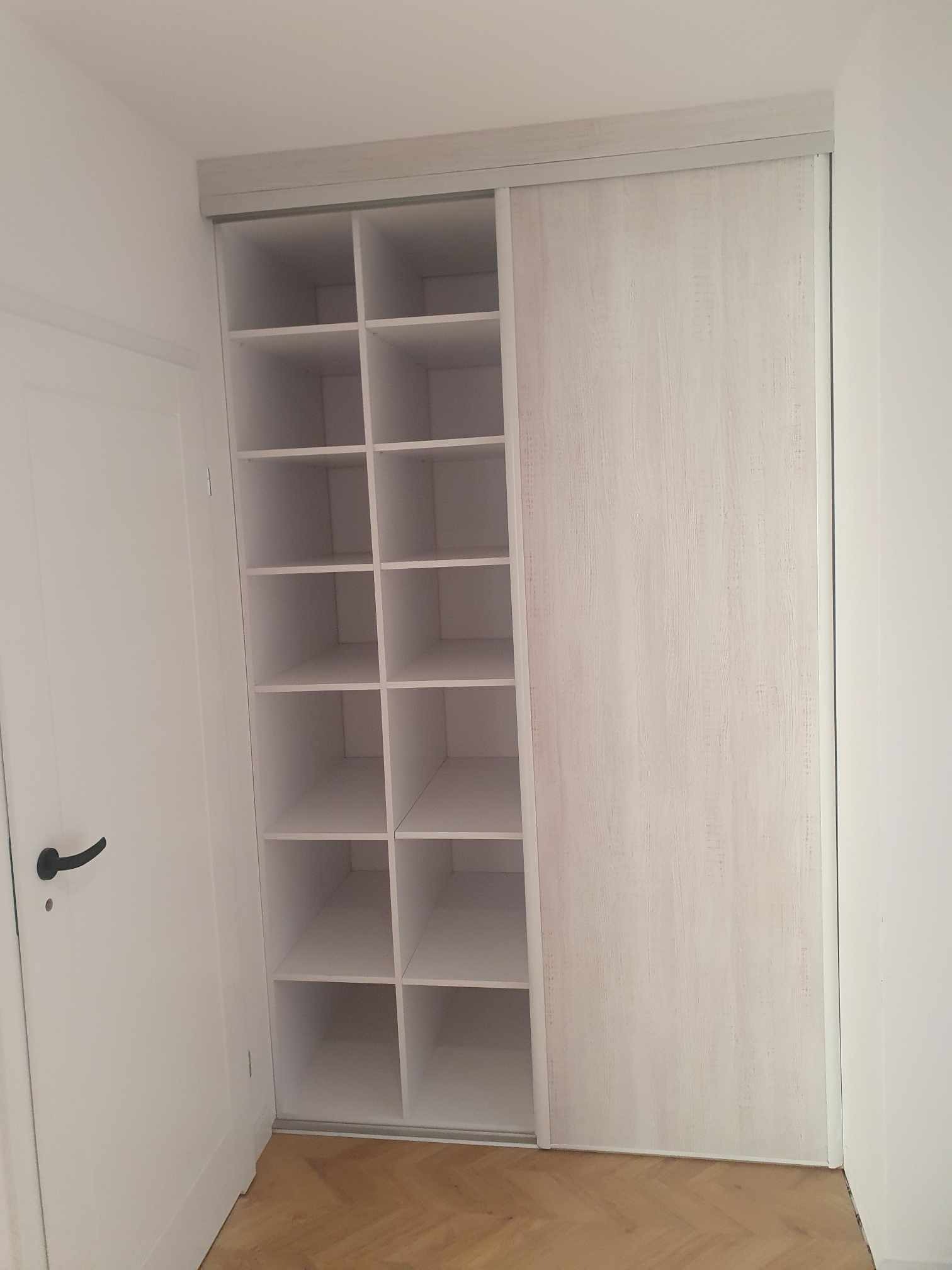 Biała szafa wnękowa z przesuwnymi drzwiami i otwartymi półkami, widok z bliska, podłoga z parkietu w jodełkę, drzwi w tle.