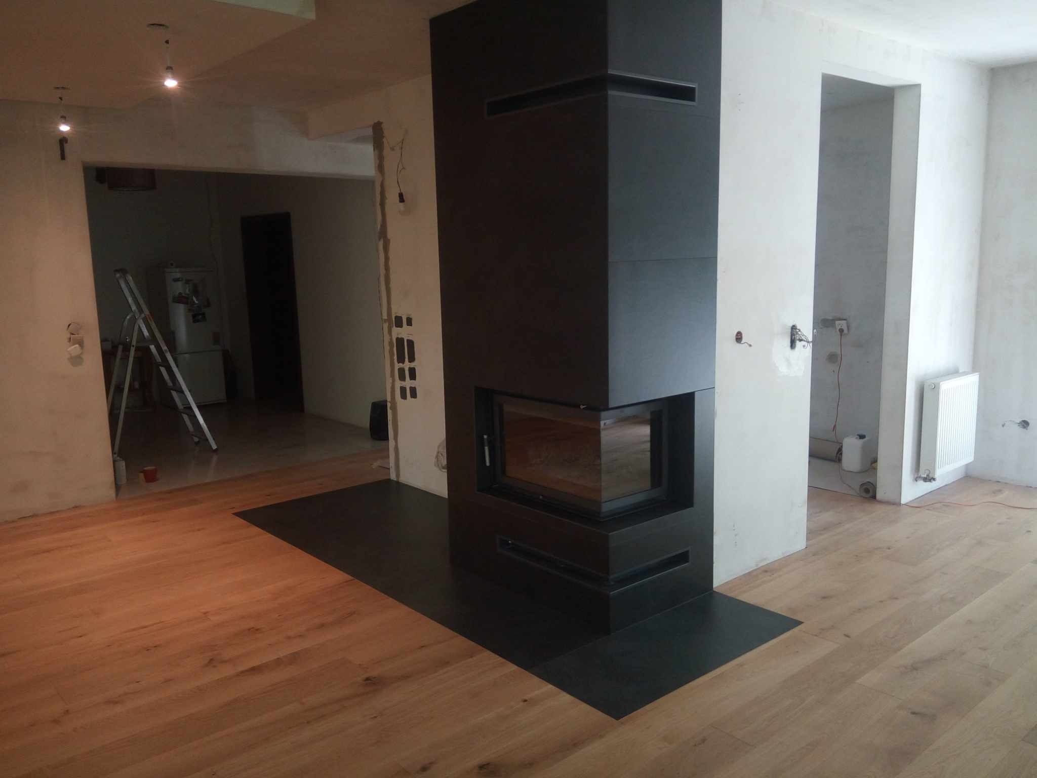 Ciemny, nowoczesny kominek w surowym wnętrzu z drewnianą podłogą. Widoczne ślady po remoncie, surowe ściany i drabina w tle. Ujęcie z niskiej perspektywy.