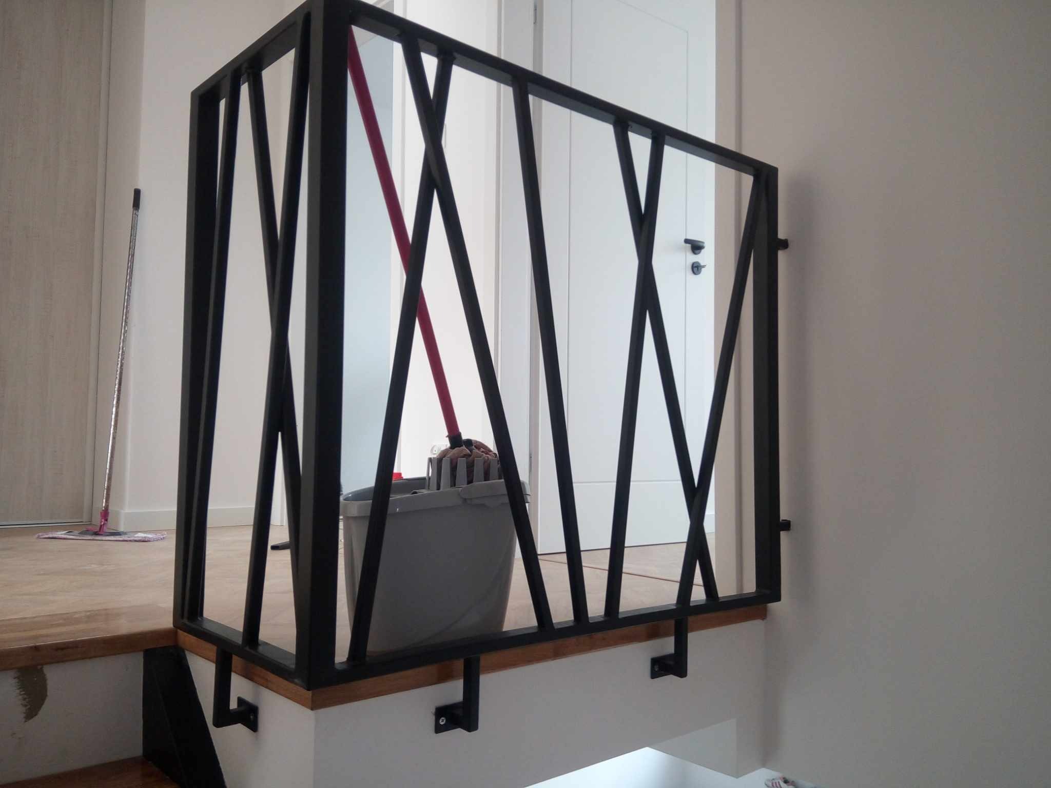 Nowoczesna balustrada schodowa z czarnych, geometrycznych elementów. Widoczny fragment schodów i mop w wiadrze w tle. Minimalistyczny design.