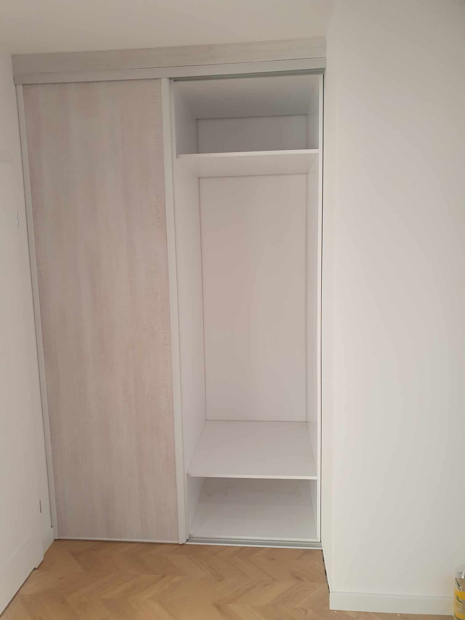 Wnękowa szafa z przesuwnymi drzwiami z delikatnym wzorem drewna, częściowo otwarta, ukazująca białe półki. Podłoga wyłożona parkietem w jodełkę.