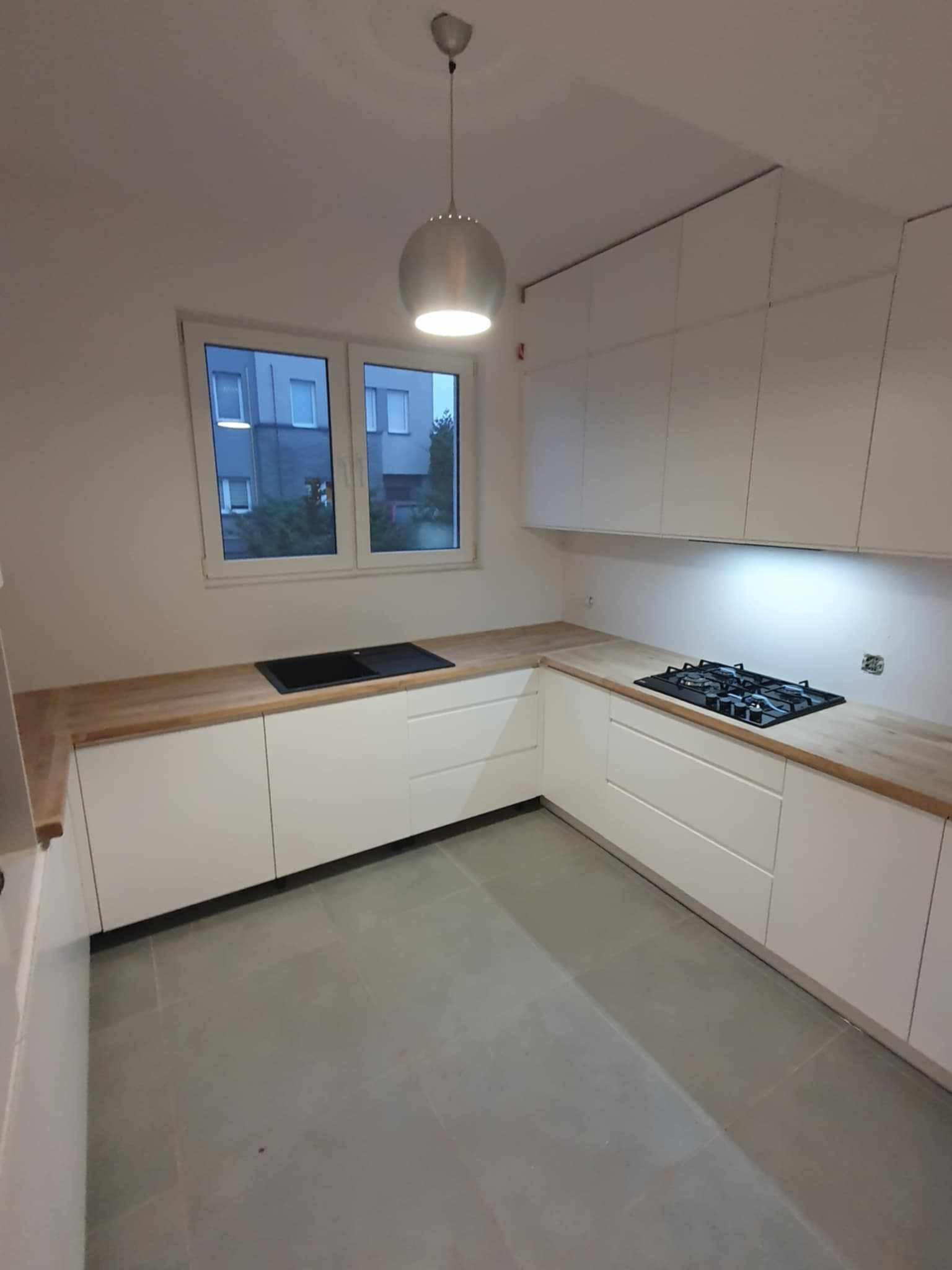 Nowoczesna kuchnia z białymi szafkami, drewnianym blatem i szarą podłogą. Widok na okno z zabudową miejską. Minimalistyczny design z oświetleniem punktowym.