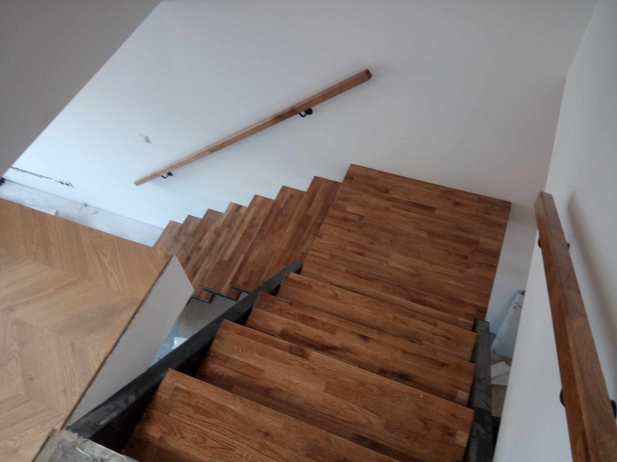 Drewniane schody z parkietem ułożonym w jodełkę na podeście. Widoczne metalowe elementy konstrukcyjne i drewniana poręcz zamontowana na ścianie.