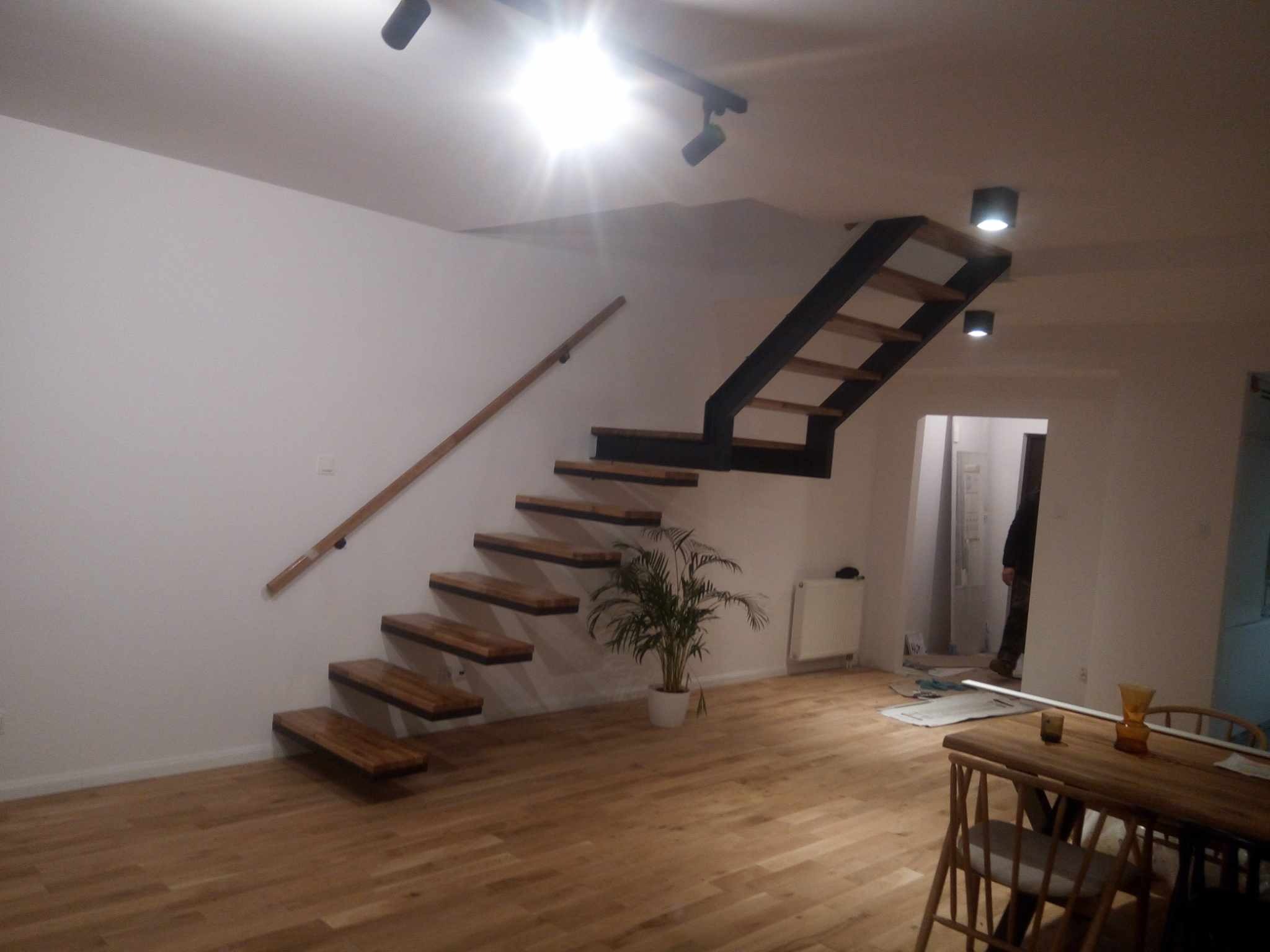 Nowoczesne, drewniane schody wiszące z metalową konstrukcją, oświetlone punktowo. Parkiet z desek w ciepłym odcieniu. Minimalistyczne wnętrze z rośliną doniczkową.