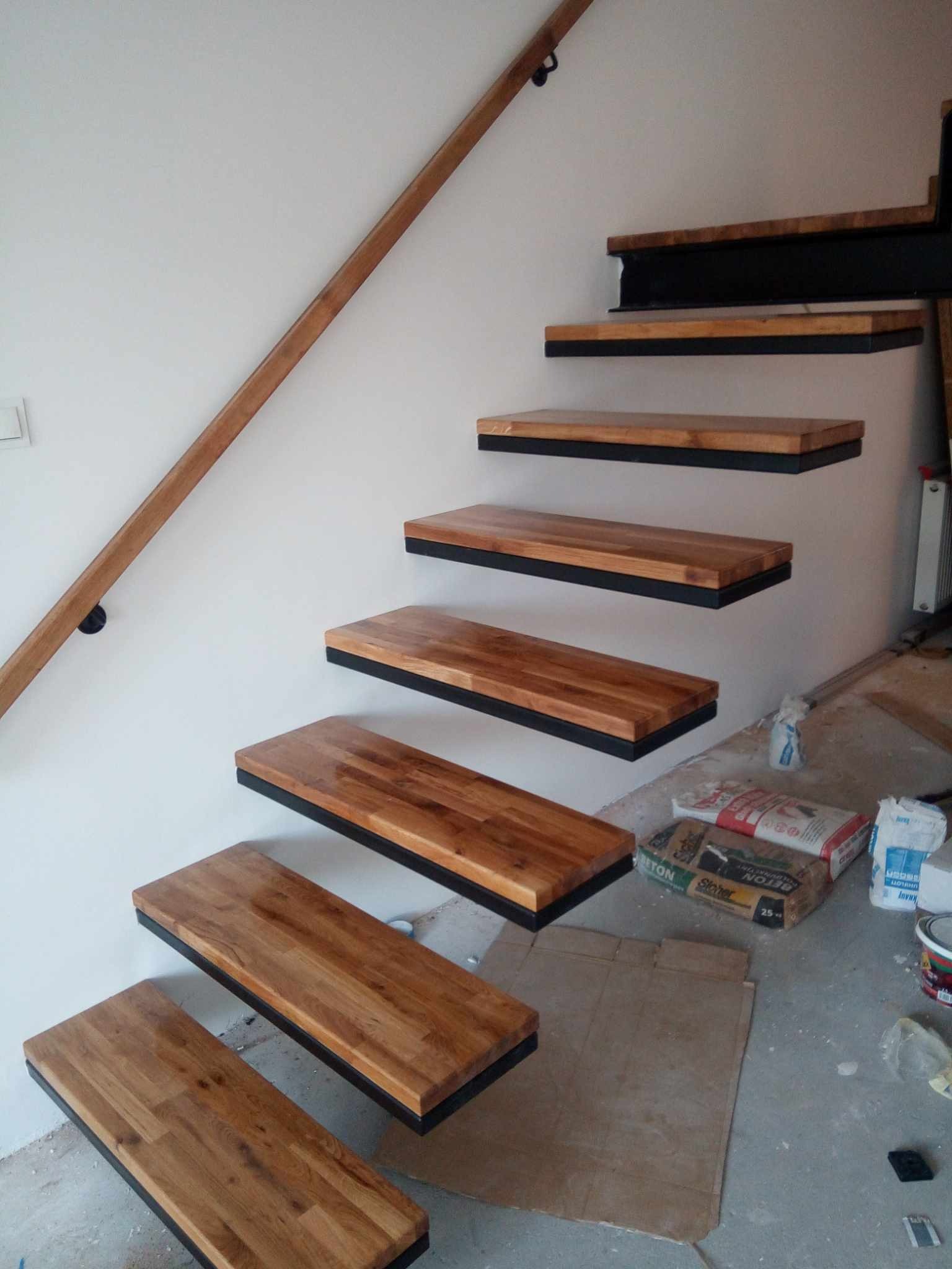 Drewniane schody wiszące z czarnymi obramowaniami, zamontowane na białej ścianie, z drewnianą poręczą. Widoczne materiały budowlane na podłodze.