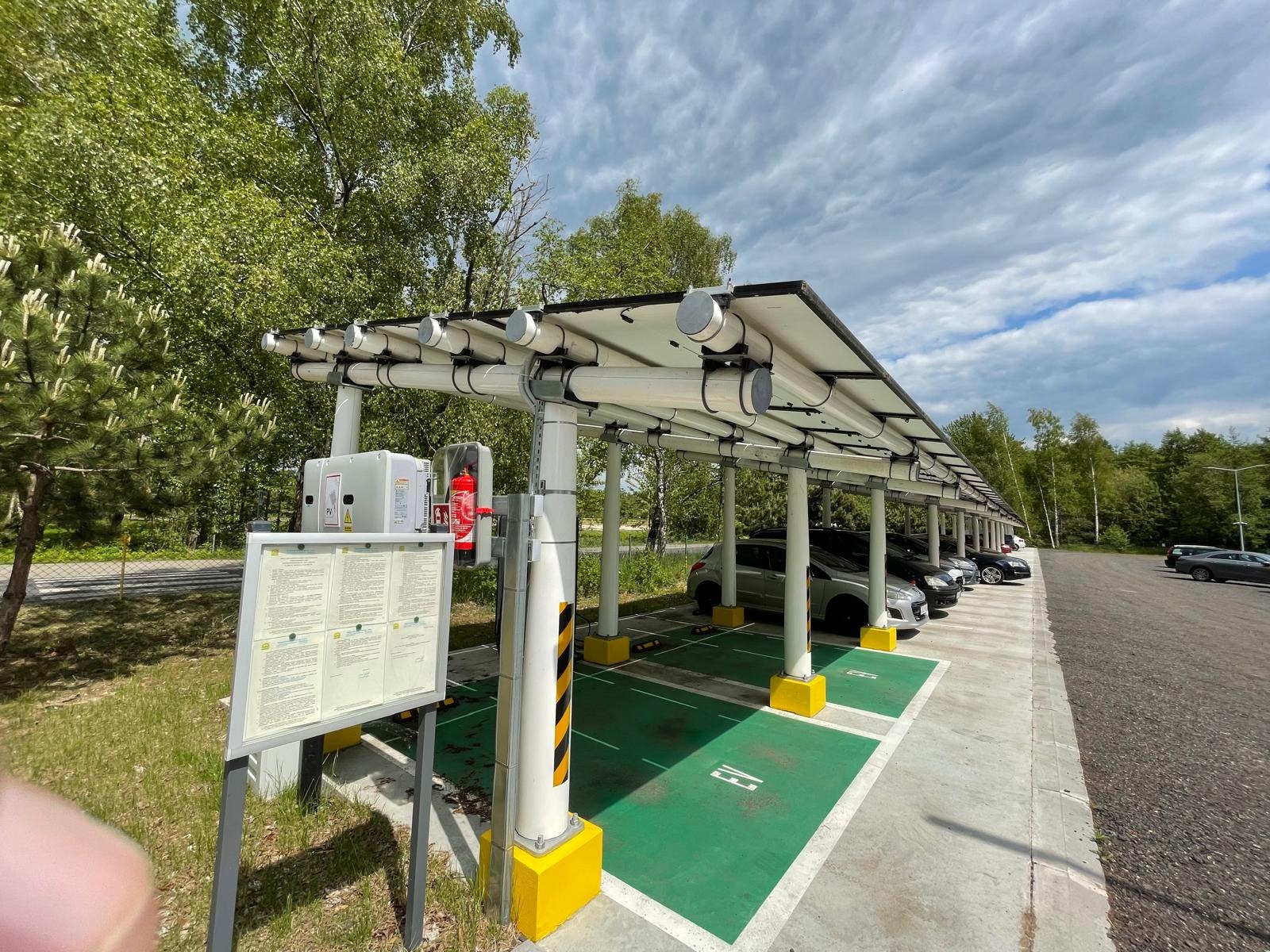 Wiaty fotowoltaiczne dla 108 samochodów w Mielnie. Kompozytowa konstrukcja z rur, z zielonymi stanowiskami EV i żółtymi obrzeżami. Widok na rząd zaparkowanych aut.