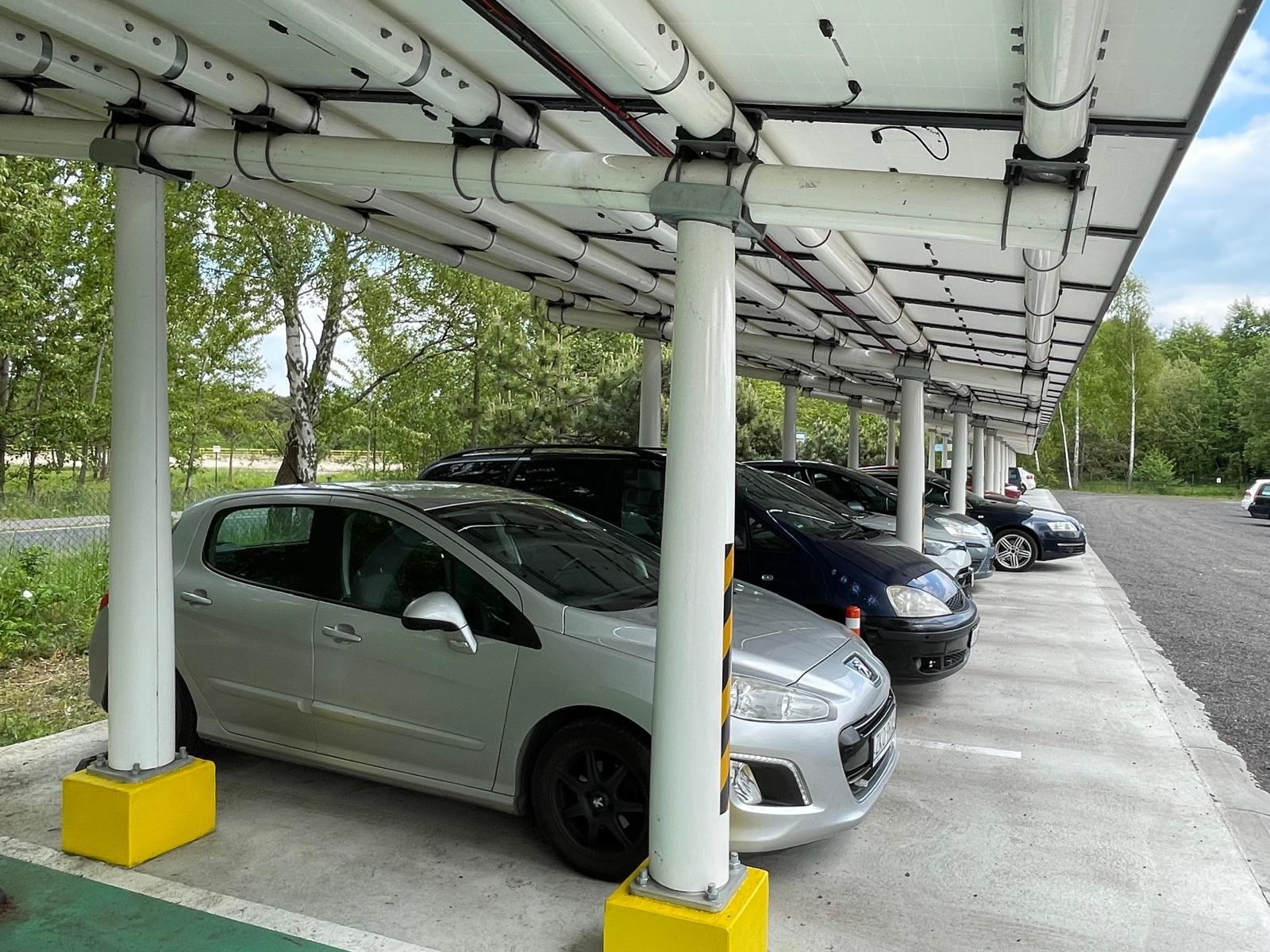 Carport fotowoltaiczny z kompozytową konstrukcją, rzędy samochodów parkują pod zadaszeniem z paneli słonecznych, wspartym na białych słupach z żółtymi podstawami.