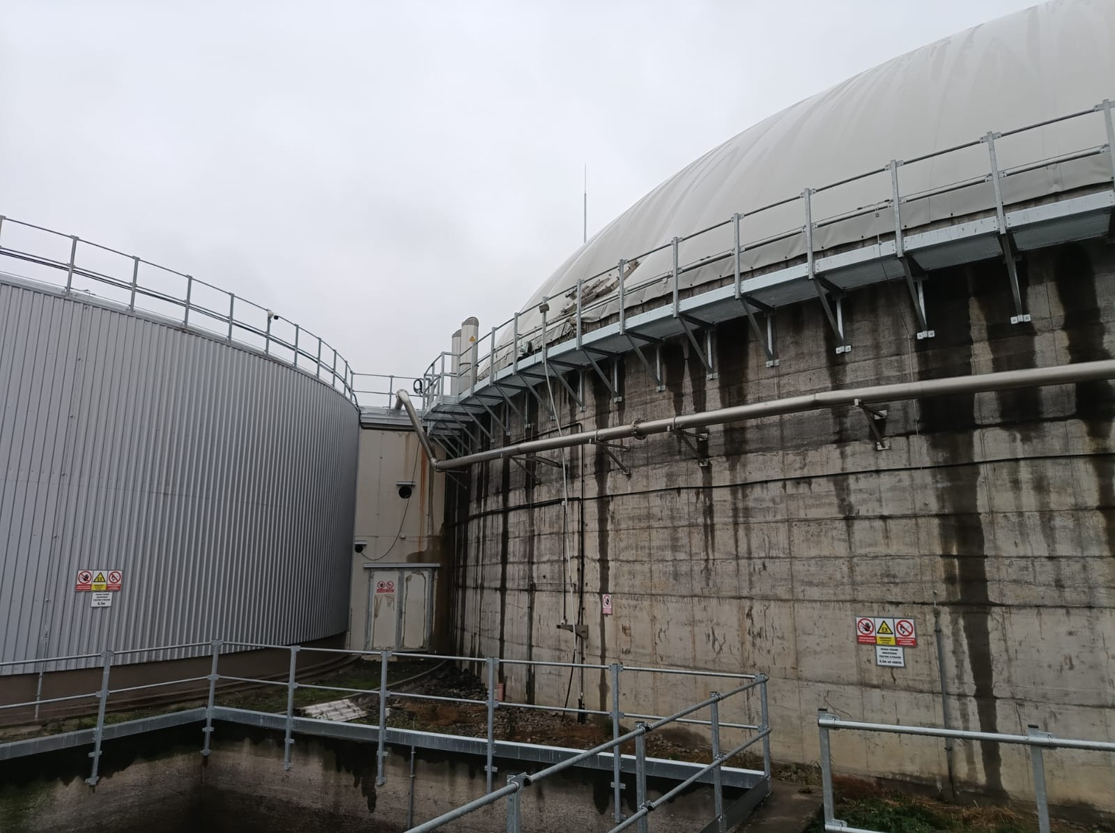 Zewnętrzny widok biogazowni z metalowymi zbiornikami, pomostami i rurami na tle pochmurnego nieba. Widoczne oznakowania bezpieczeństwa i ślady po zaciekach na betonowej konstrukcji.