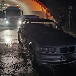 POMOC DROGOWA ŁUKASZ PRZYBORSKI - Uszkodzone auto BMW z otwartą maską, holowane przez lawetę w tunelu. Mokra nawierzchnia, nocne oświetlenie, widoczny znak drogowy.